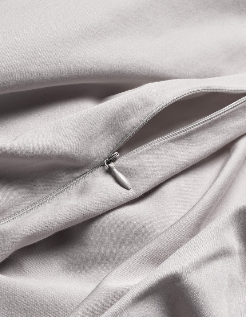 A close-up of Cozy Earth's Soft-Wash Cotton Duvet Cover in light grey shows a silver zipper partially unzipped on its smooth, gently folded fabric.