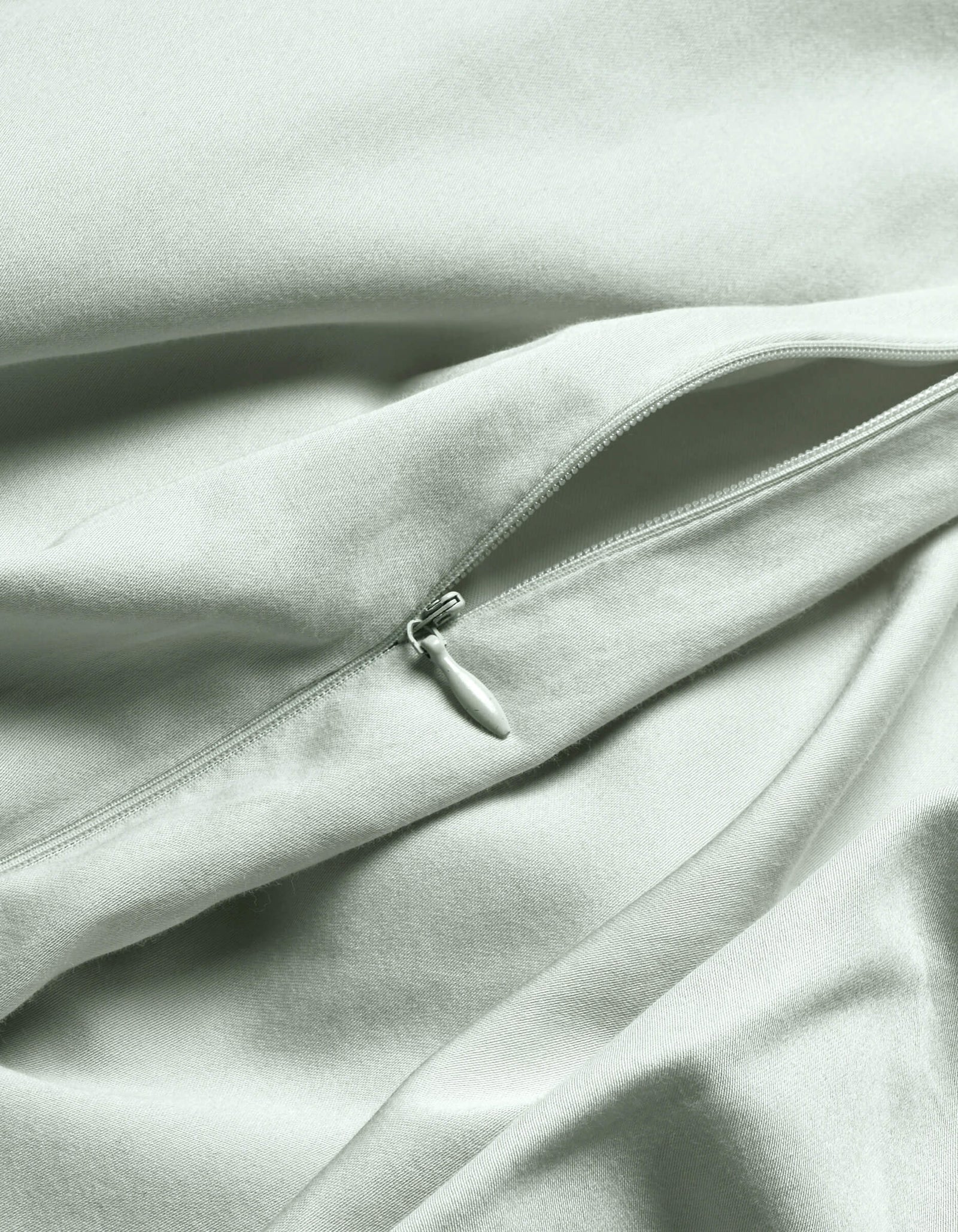 A close-up of the Cozy Earth Soft-Wash Cotton Duvet Cover in light gray or pale green shows a visible, partially unzipped zipper creating gentle folds and soft shadows on the smooth fabric.