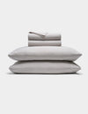 A neatly folded Soft-Wash Cotton Sheet Set by Cozy Earth in light gray is stacked atop two matching pillows, all set against a plain white background.|Color:Dew