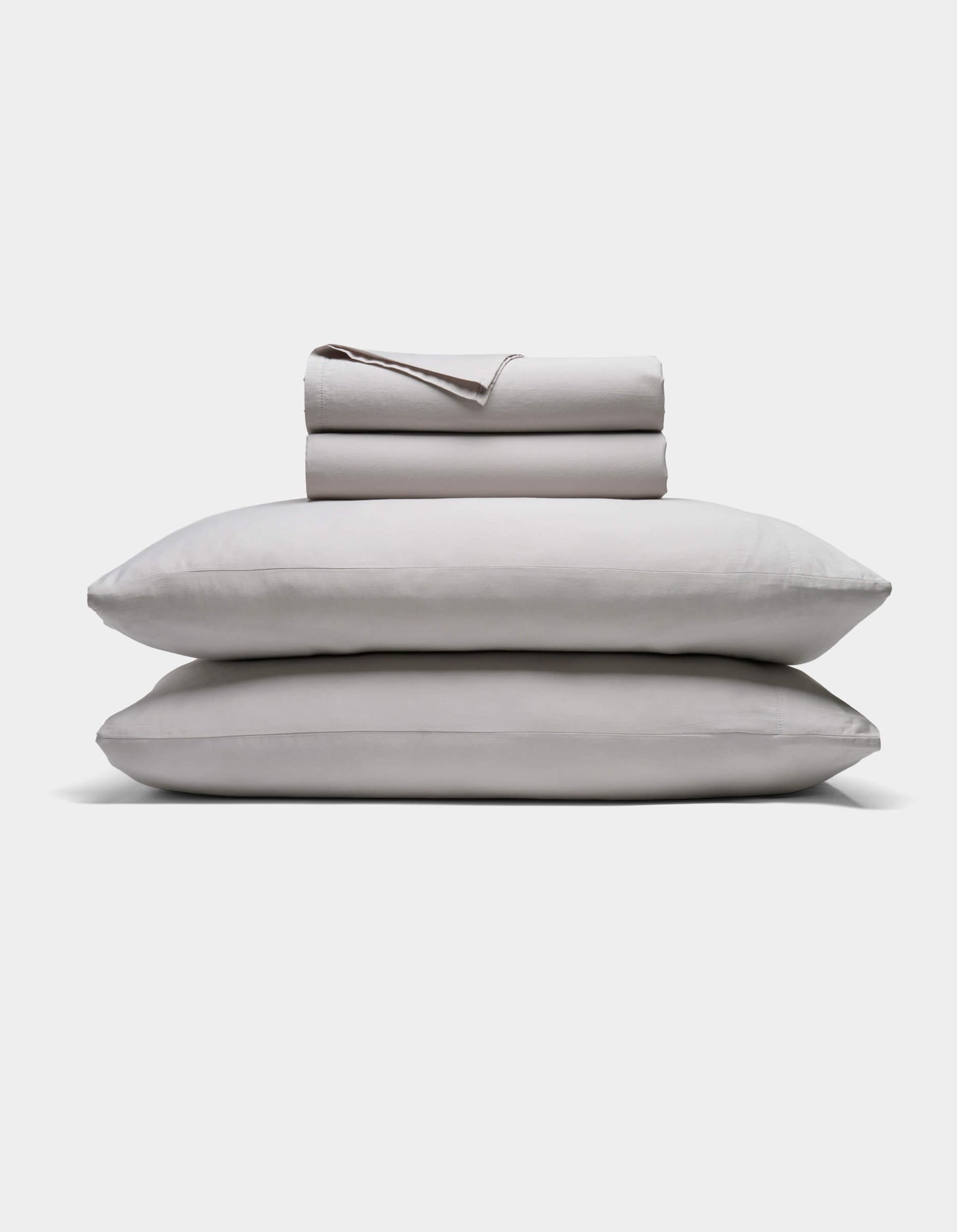 A neatly folded Soft-Wash Cotton Sheet Set by Cozy Earth in light gray is stacked atop two matching pillows, all set against a plain white background.