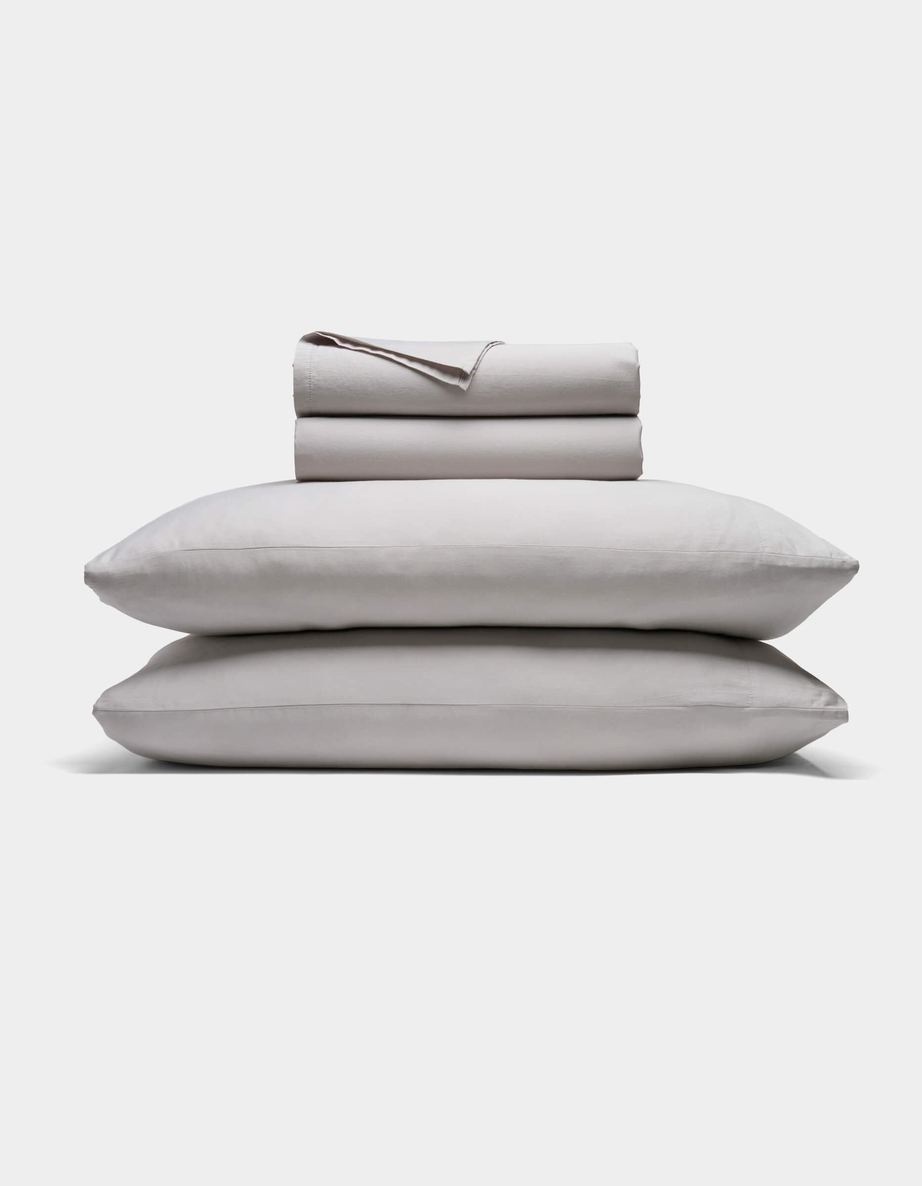 A neatly folded Soft-Wash Cotton Sheet Set by Cozy Earth in light gray is stacked atop two matching pillows, all set against a plain white background.|Color:Dew