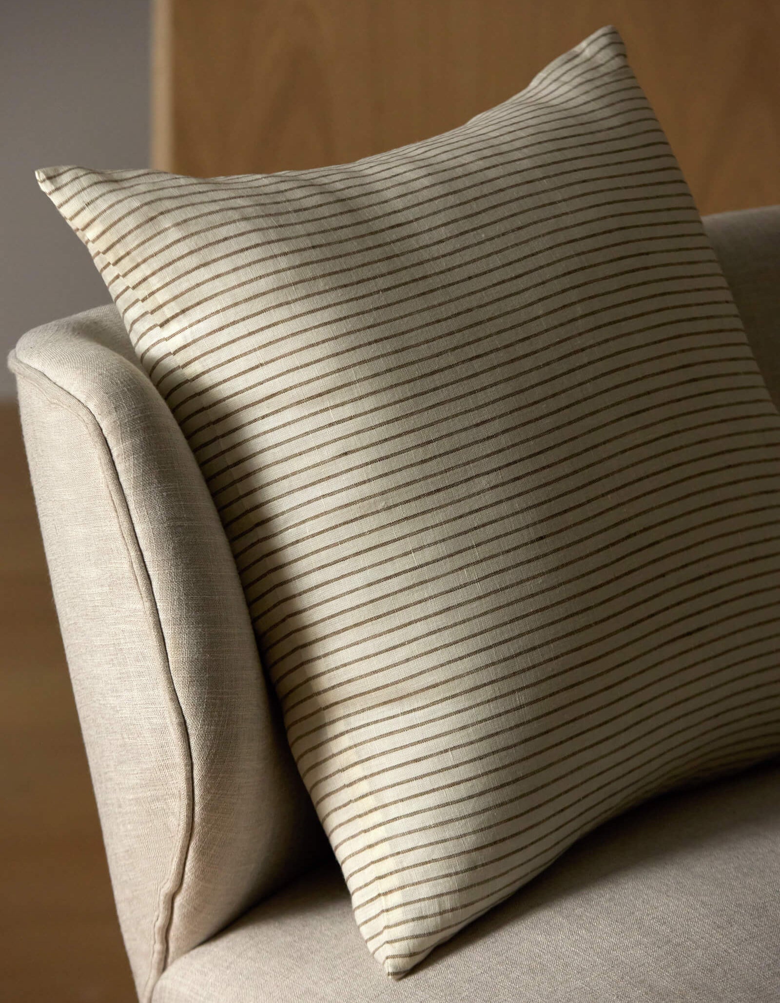 The Striped Throw Pillow by Cozy Earth, featuring thin horizontal brown stripes on beige, rests on a light upholstered chair with a wooden panel in the background.