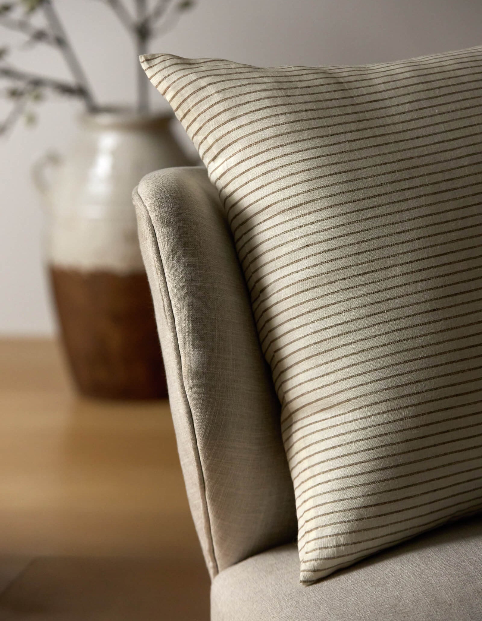 A close-up of the Cozy Earth Striped Throw Pillow resting on a light armchair, with a blurred ceramic vase and branches in the background.