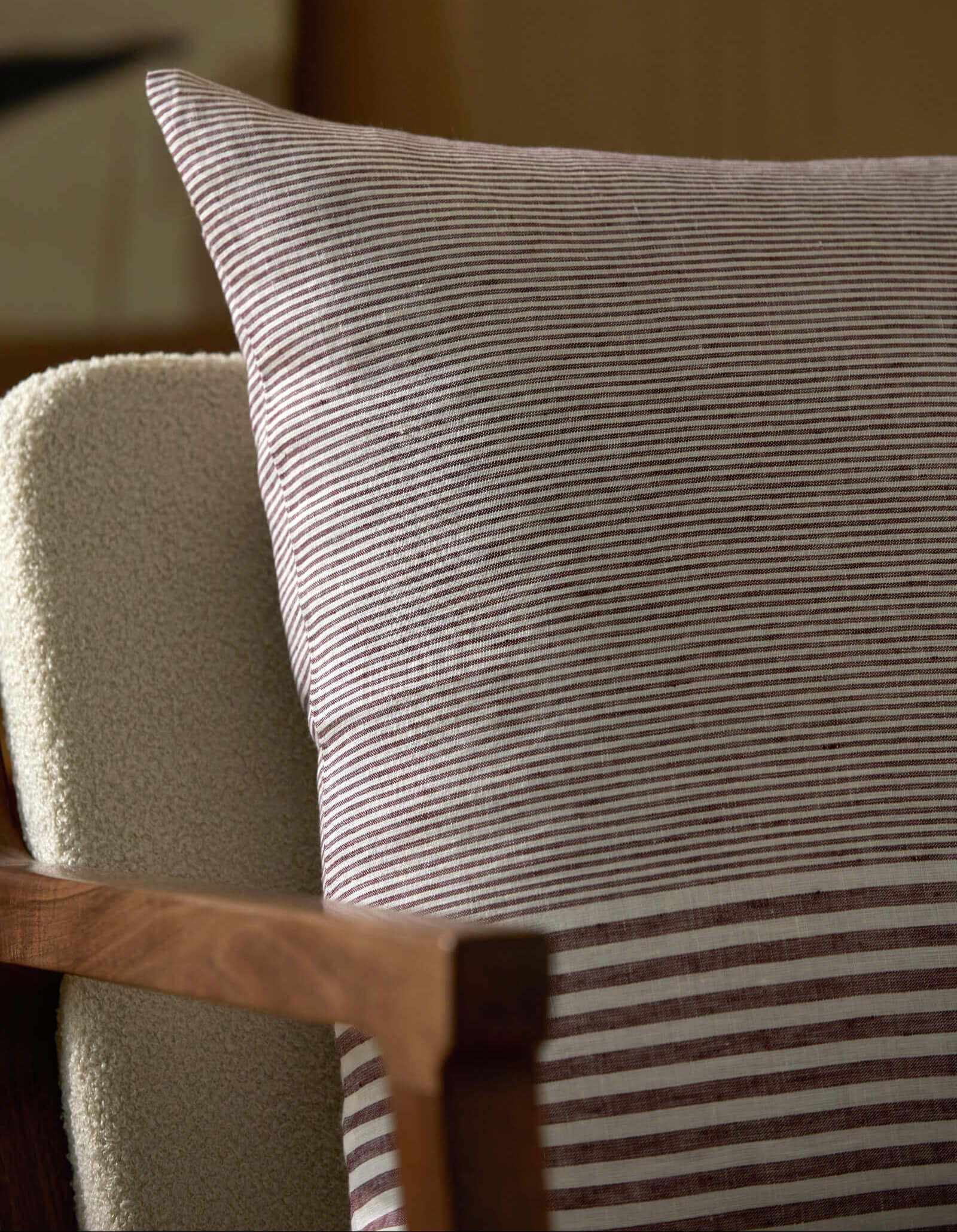 The Cozy Earth Striped Throw Pillow, featuring thin dark lines, rests on a beige textured armchair with a wooden frame. The softly blurred background highlights the pillow’s elegant design.