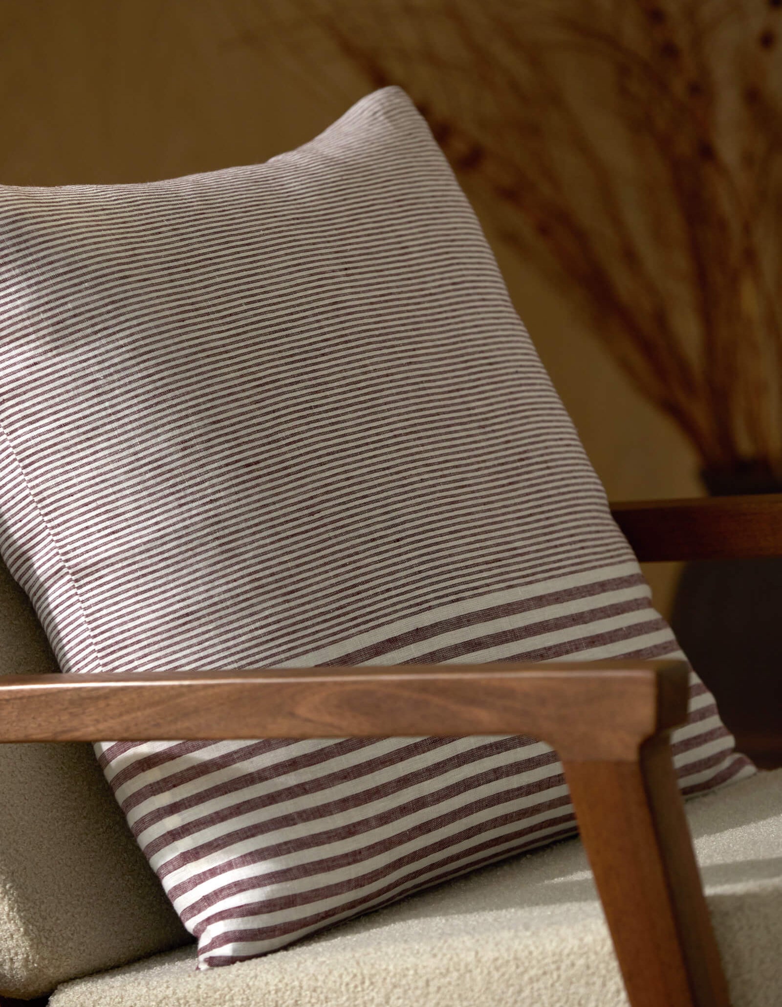 A close-up of a beige armchair with wooden armrests showcases the Cozy Earth Striped Throw Pillow in gray and white. Blurred dried branches in a vase are visible in the background.