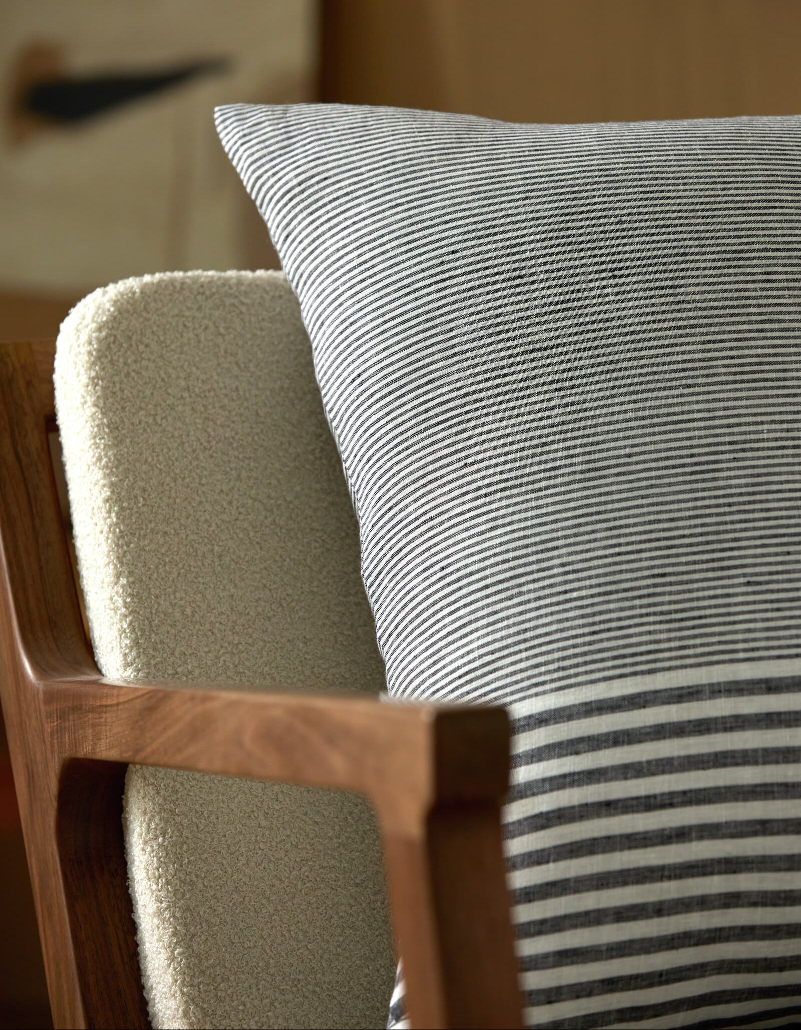 A close-up of a wooden chair features a textured cream cushion and Cozy Earth’s Striped Throw Pillow in gray and white resting against the backrest, with a softly blurred background hinting at a cozy indoor setting.