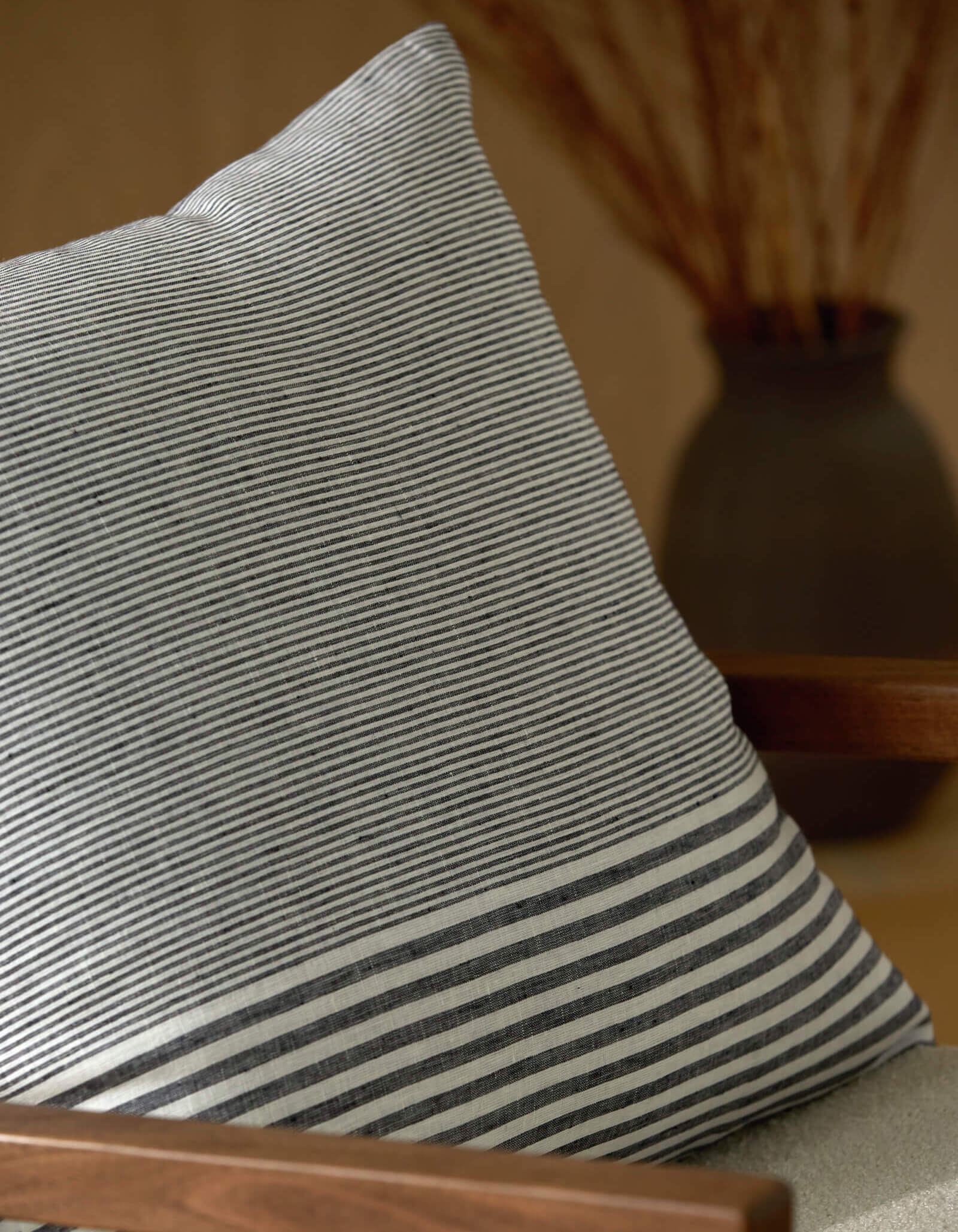 A close-up of the Cozy Earth Striped Throw Pillow, featuring thin black and white stripes, rests on a wooden chair. In the blurred background, a dark vase with tall branches adds contrast.
