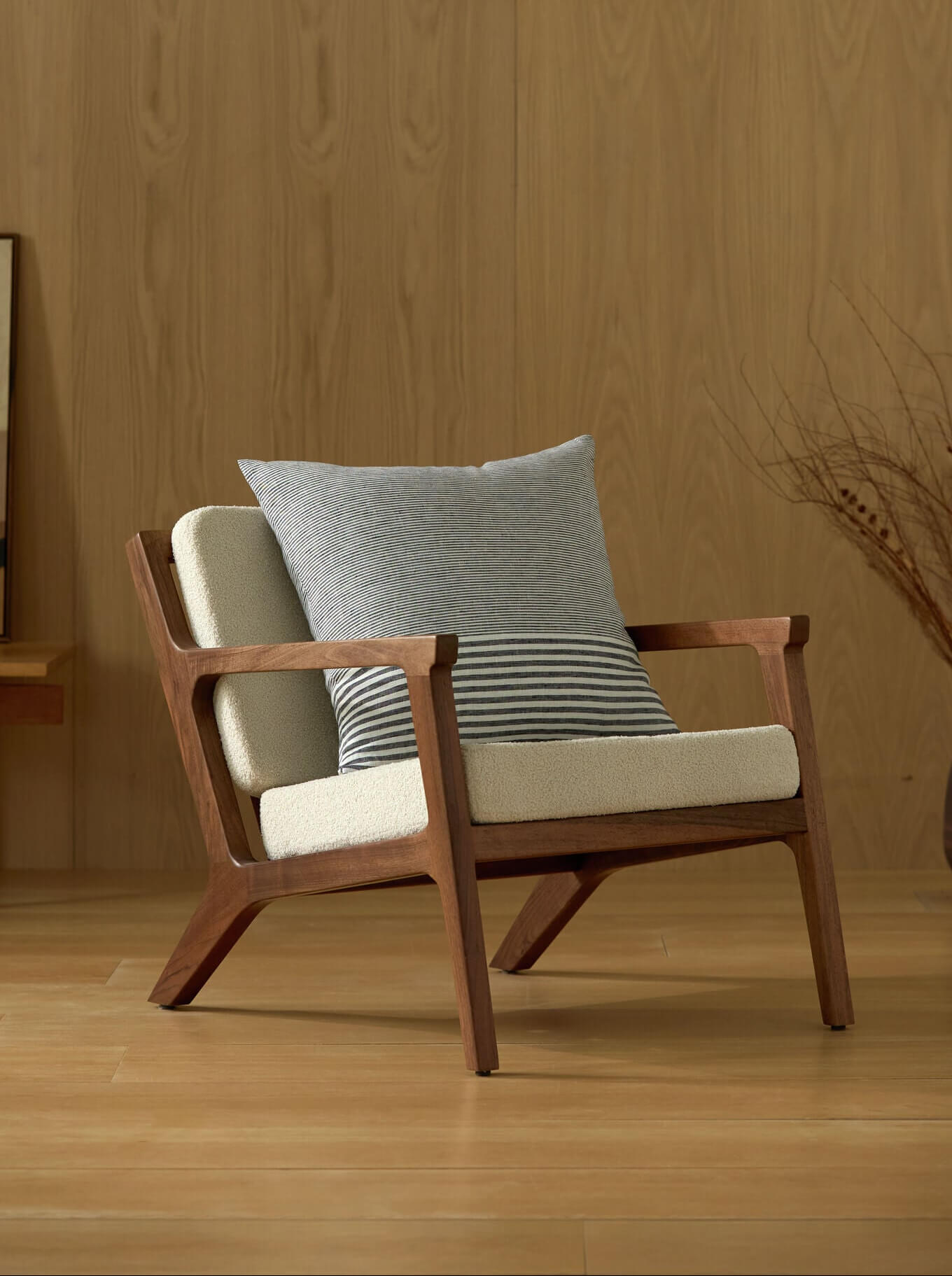 A modern wooden armchair with cream cushions and a Cozy Earth Striped Throw Pillow sits on a wooden floor before a light wood-paneled wall, with abstract art and a vase of dried branches in the background.