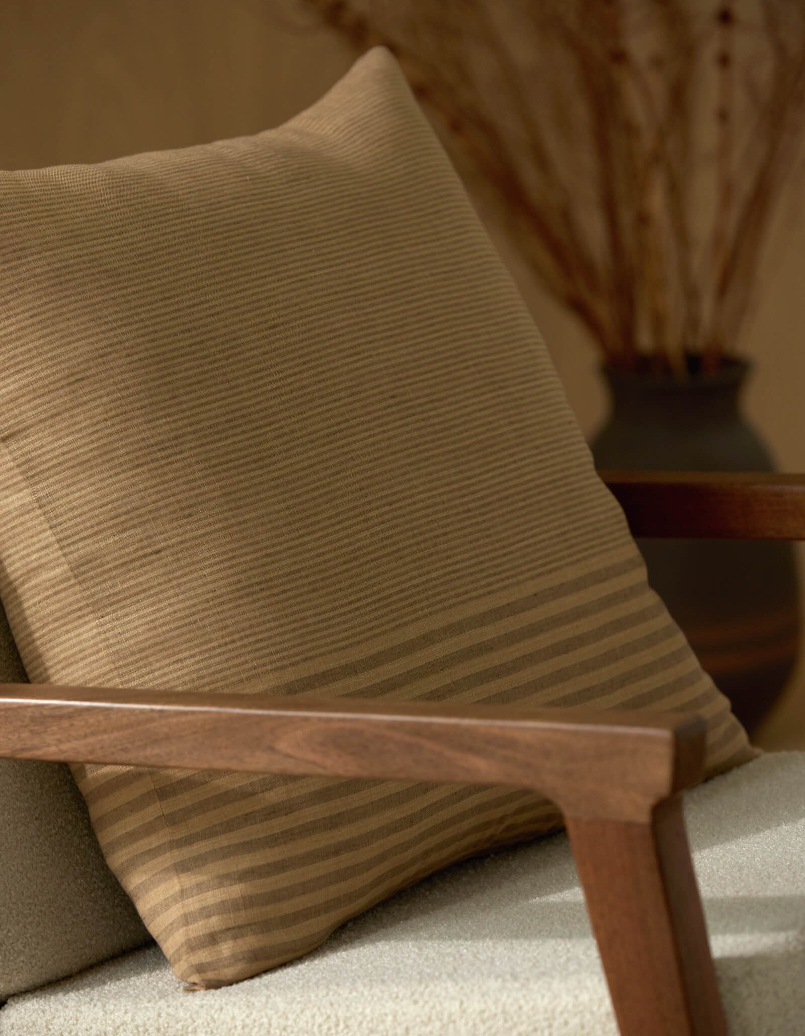 A close-up of a wooden armchair featuring the Cozy Earth Striped Throw Pillow, with a blurred vase of dried branches in the background.