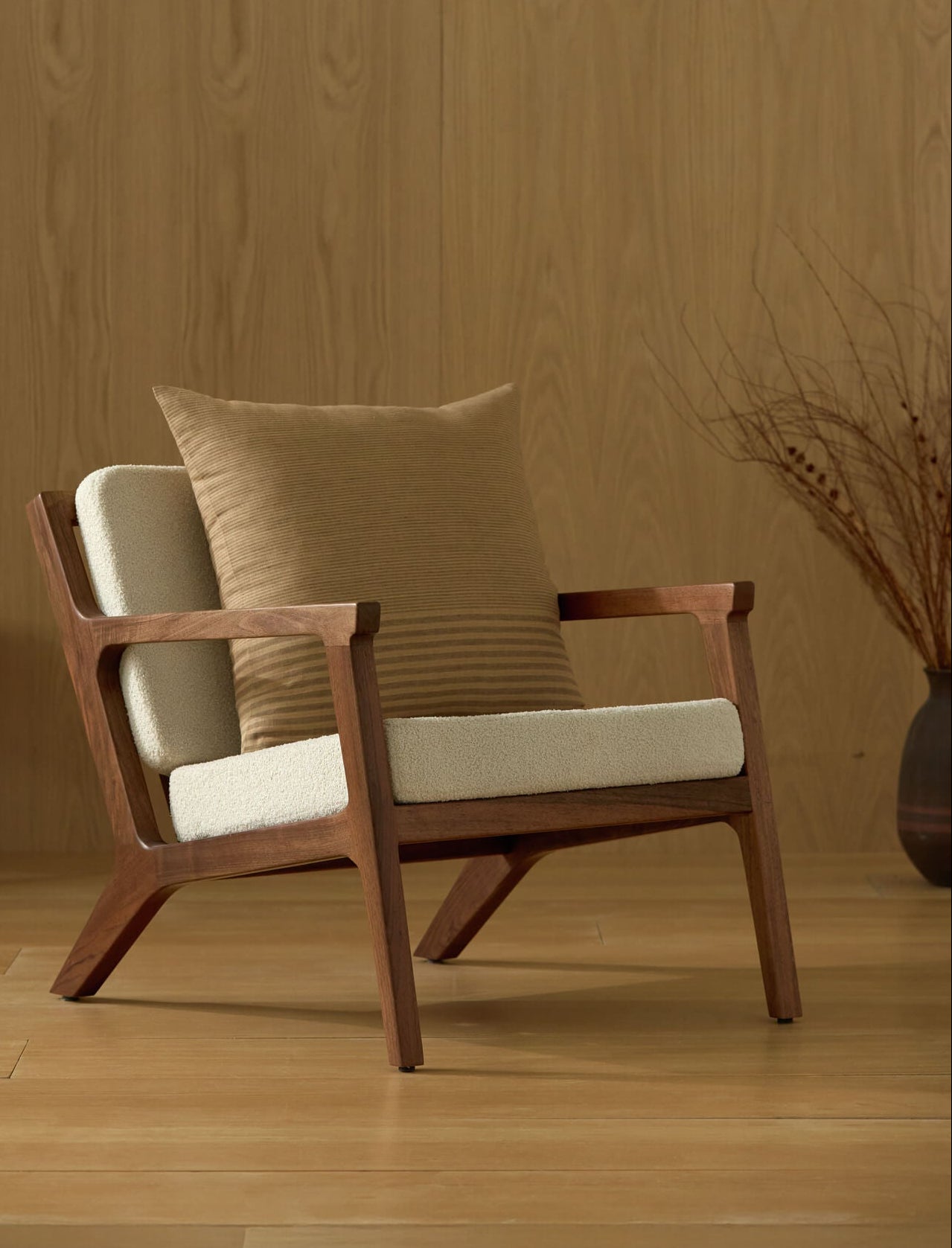 A modern wooden armchair with cream cushions and a Cozy Earth Striped Throw Pillow sits on a light wood floor. A minimalist painting leans against a wood-paneled wall, with dried branches in a vase beside the chair.