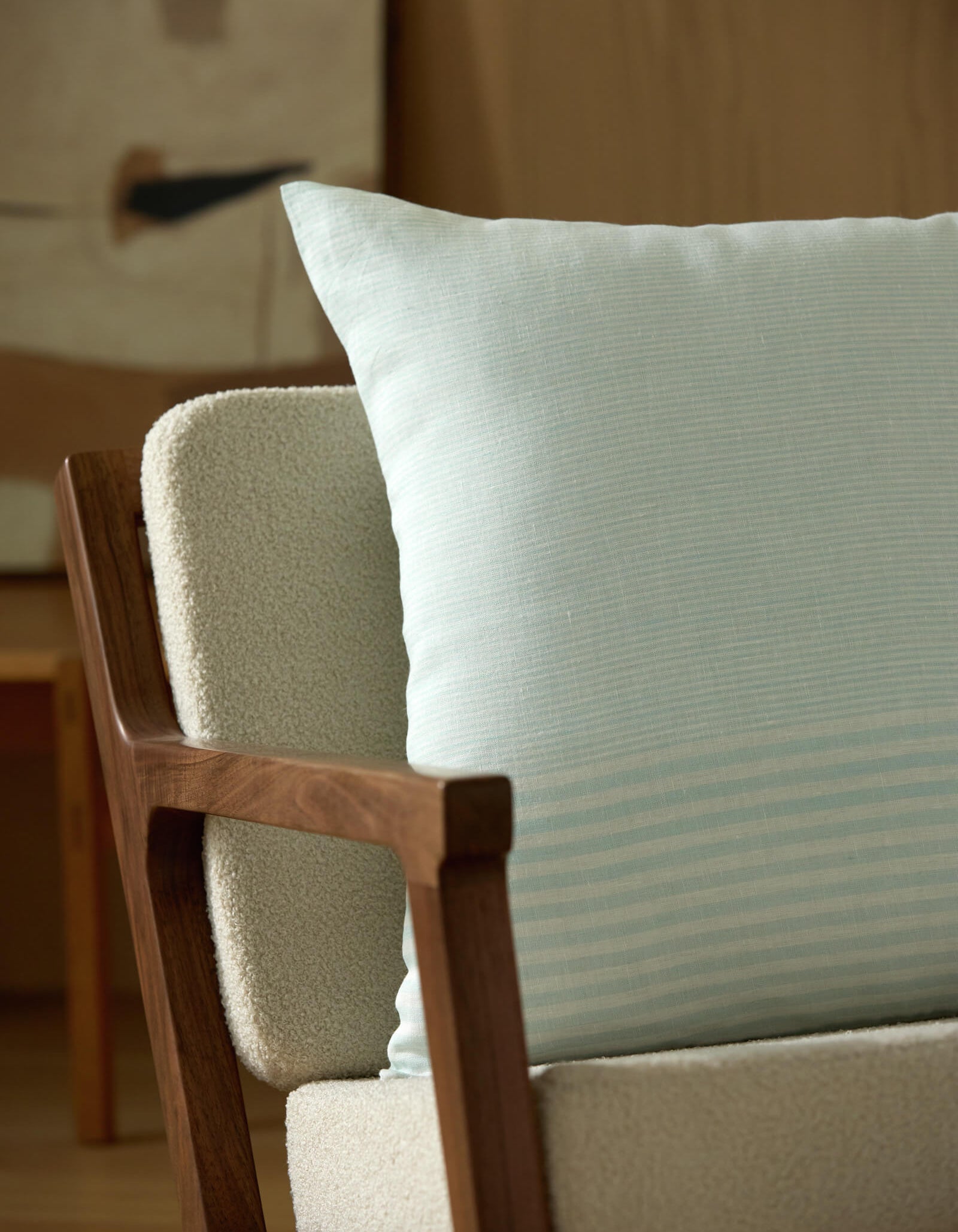 A close-up of a modern armchair with wooden armrests, light textured upholstery, and a Striped Throw Pillow by Cozy Earth. An abstract painting and wooden furniture are softly blurred in the background.