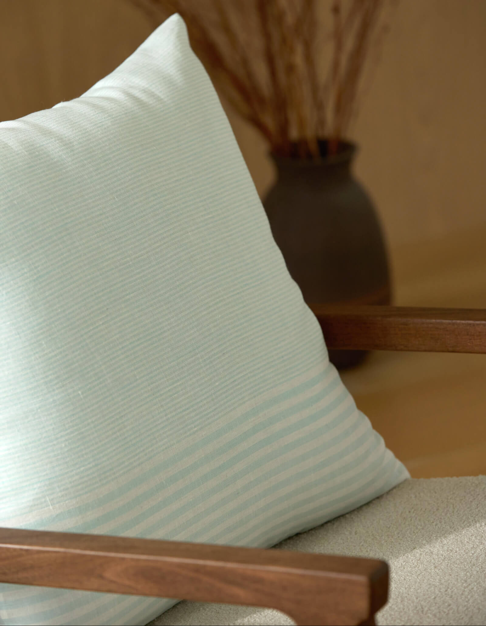 The Striped Throw Pillow by Cozy Earth in light blue and white sits on a wooden chair, while a dark vase with dried branches is blurred behind it. Soft lighting adds to the calm and cozy feel of the scene.