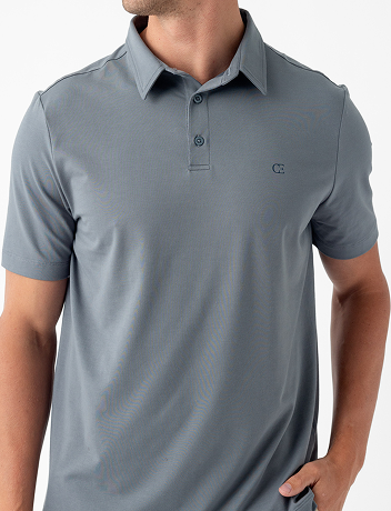 Man wearing a gray polo shirt with short sleeves. One hand is in his pocket, and he is turned slightly to the side. The shirt has a small logo on the chest and features a button-up collar. The background is plain white.