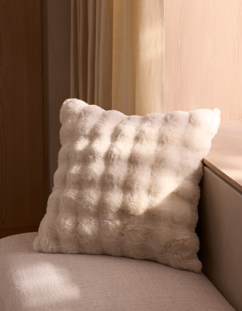 The Cozy Earth Bubble Cuddle Pillow, soft and fluffy in white, sits on a light beige seat near a wall as sunlight streams through the window, casting gentle shadows on the pillow and surrounding surfaces.