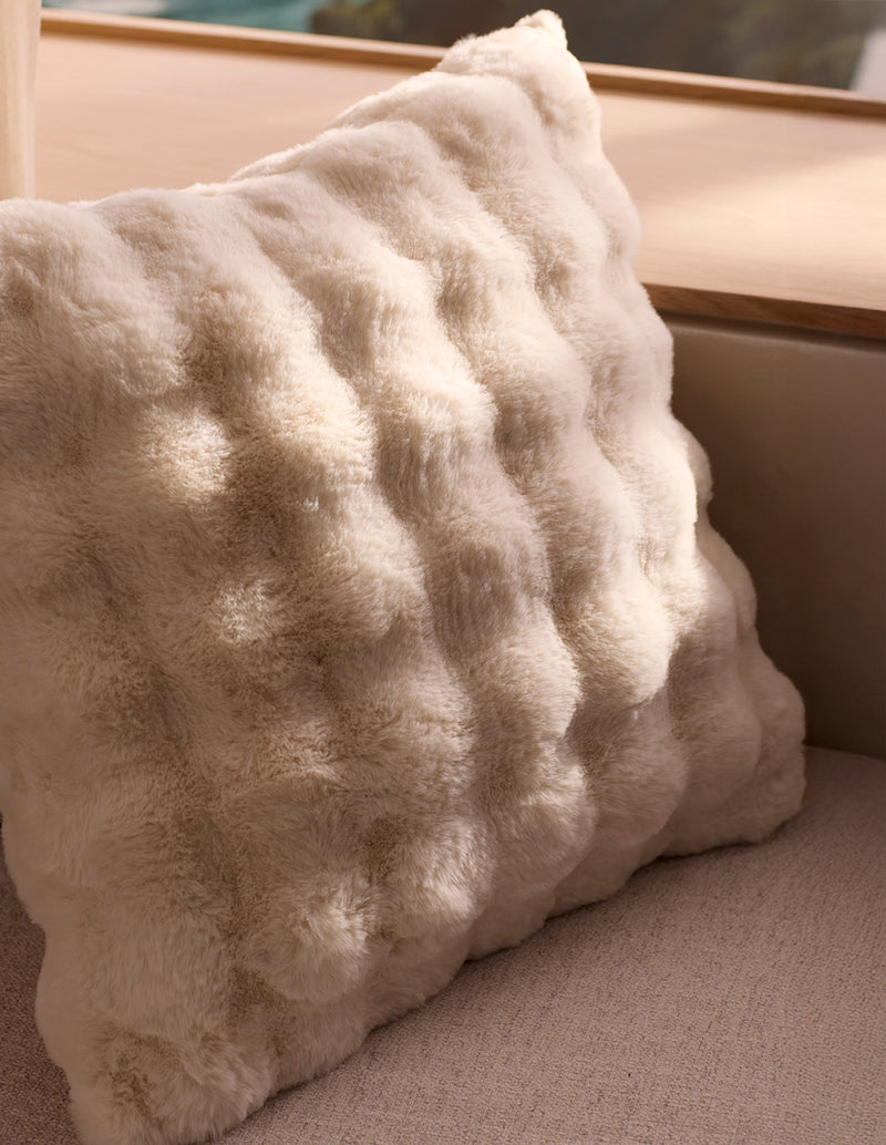 The Cozy Earth Bubble Cuddle Pillow, a soft cream faux fur with a raised texture, rests on a light beige couch by a wooden window ledge, illuminated by natural sunlight.