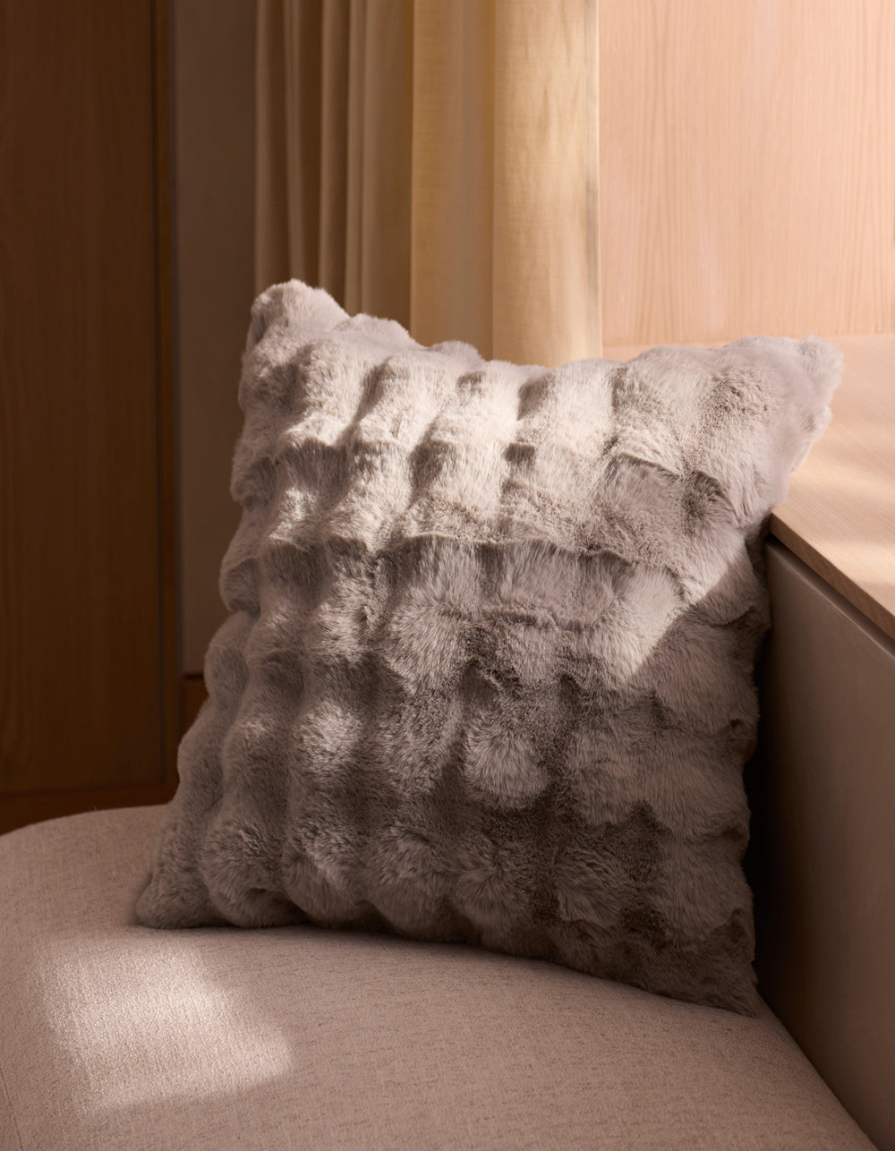 The Bubble Cuddle Pillow by Cozy Earth, featuring textured gray faux fur, rests on a light beige upholstered bench beside a wooden wall and cream curtains, with natural light casting gentle shadows.