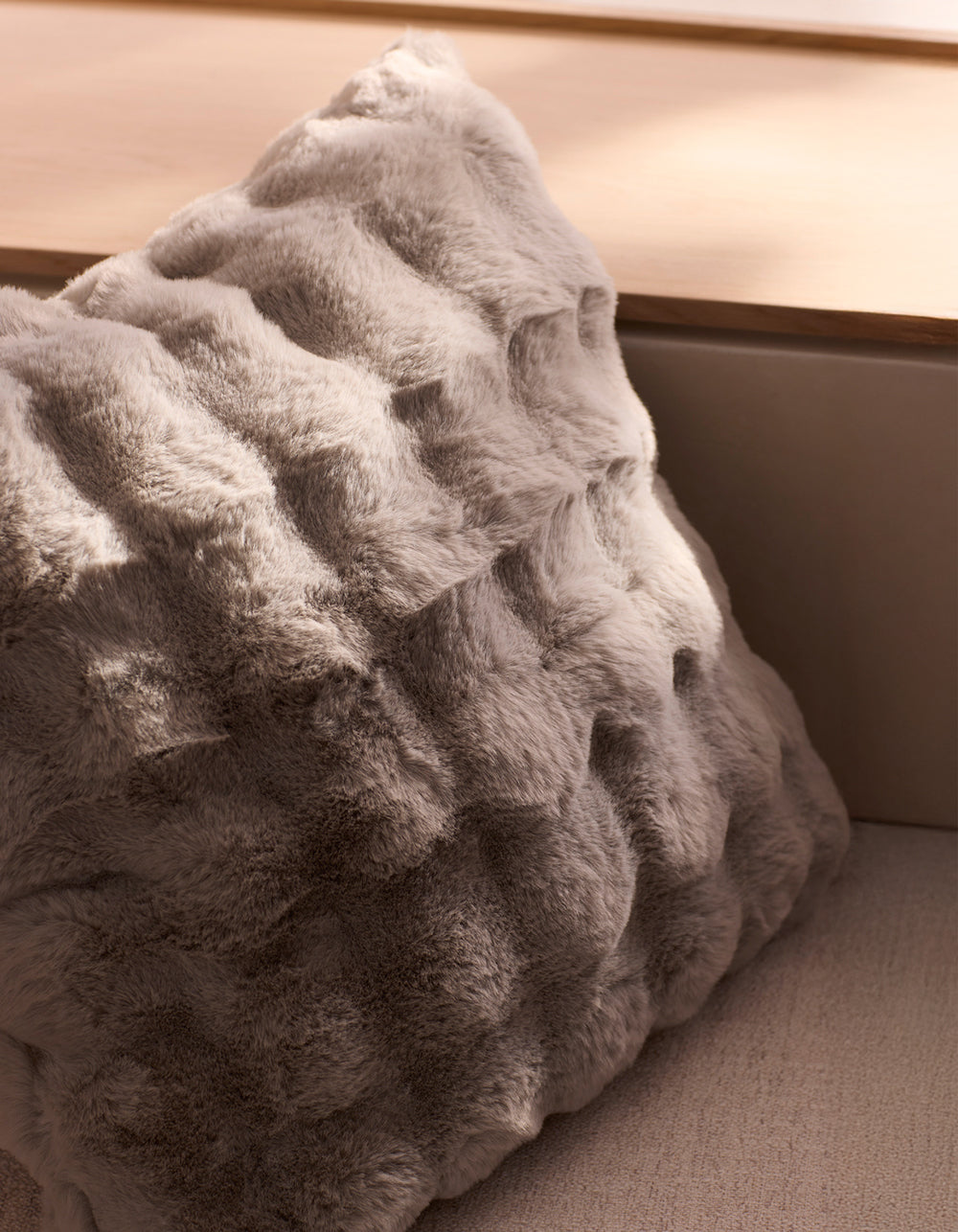 The Bubble Cuddle Pillow by Cozy Earth, in light grey, features a textured, fur-like surface gently illuminated by sunlight as it rests against a wooden ledge.
