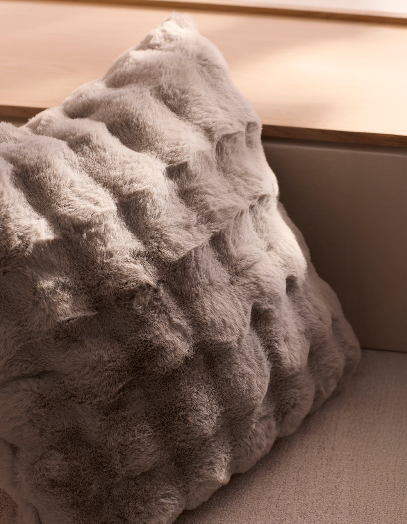 The Bubble Cuddle Pillow by Cozy Earth, featuring soft gray faux fur with a wavy texture, rests on a light wooden surface in a sunlit, neutral-toned room.|Color:Light Grey