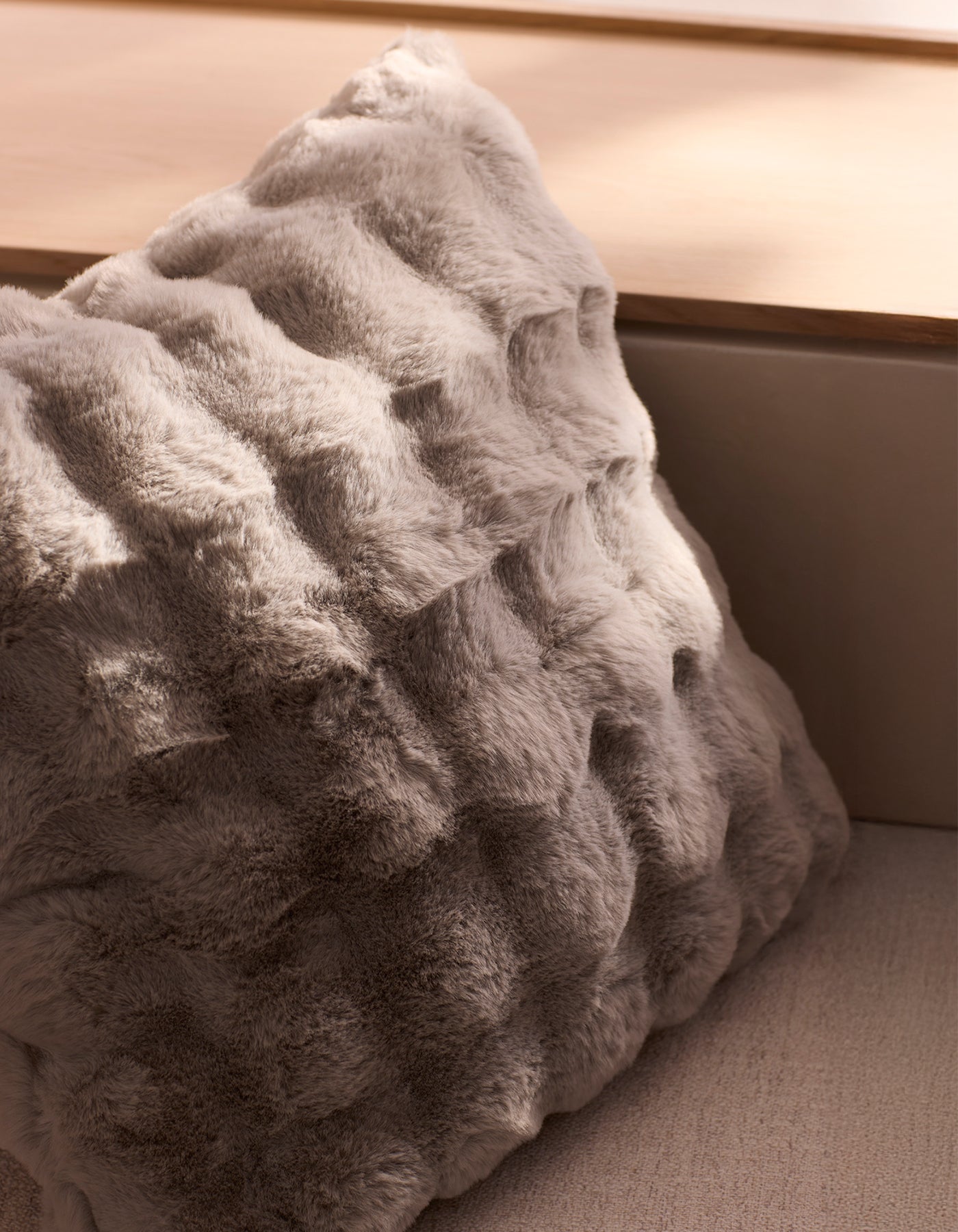 The Bubble Cuddle Pillow by Cozy Earth, featuring soft gray faux fur with a wavy texture, rests on a light wooden surface in a sunlit, neutral-toned room.