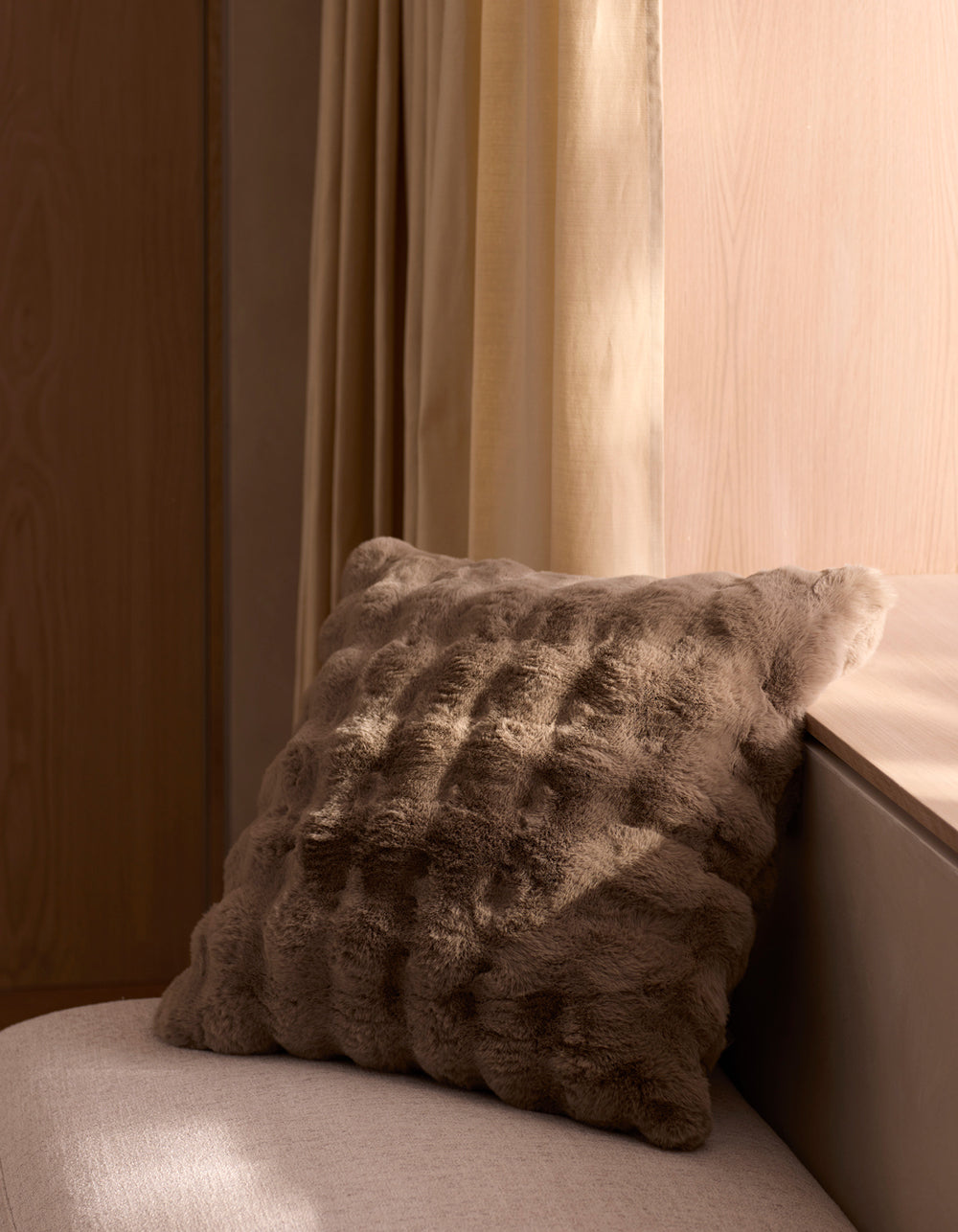 The Cozy Earth Bubble Cuddle Pillow sits on a light beige cushioned bench by pale wood walls and a window with cream curtains, softly illuminated by natural light.