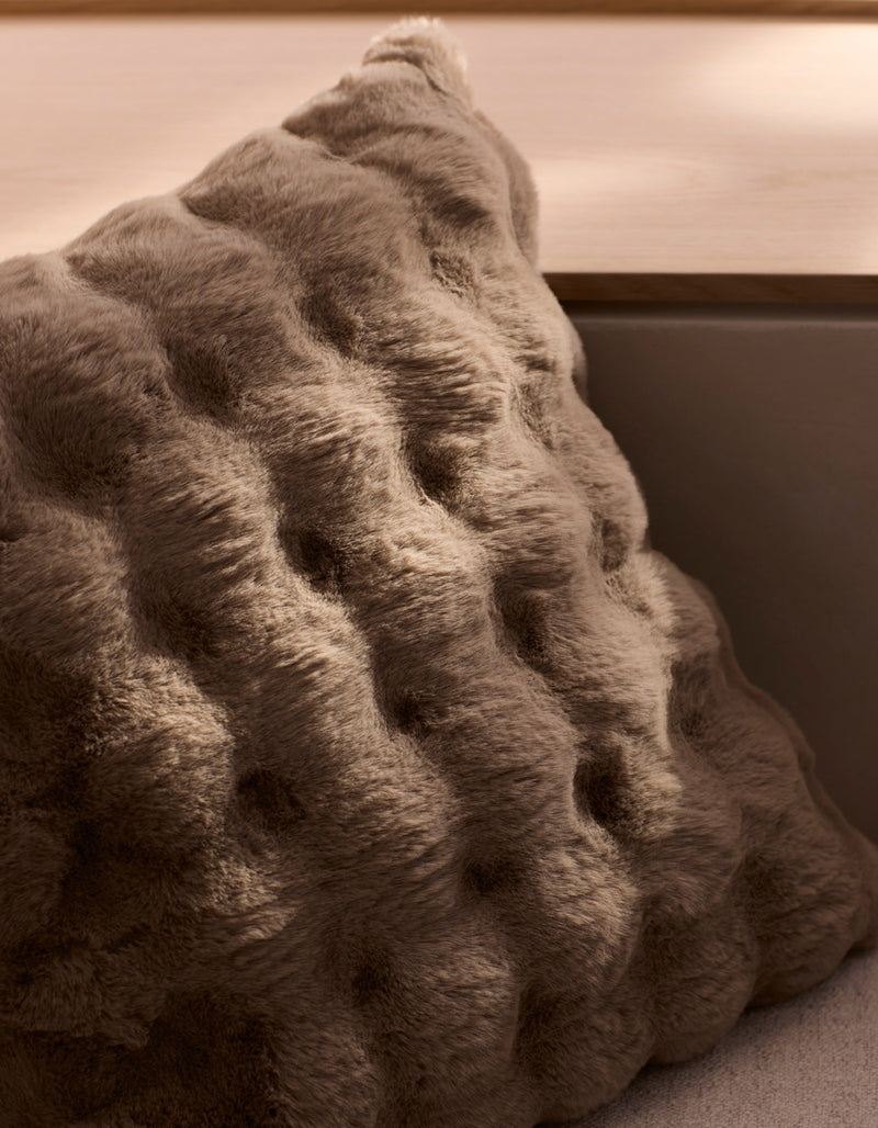 The Cozy Earth Bubble Cuddle Pillow, with its plush brown wavy tufted texture, rests on a light wooden bench in soft natural light.