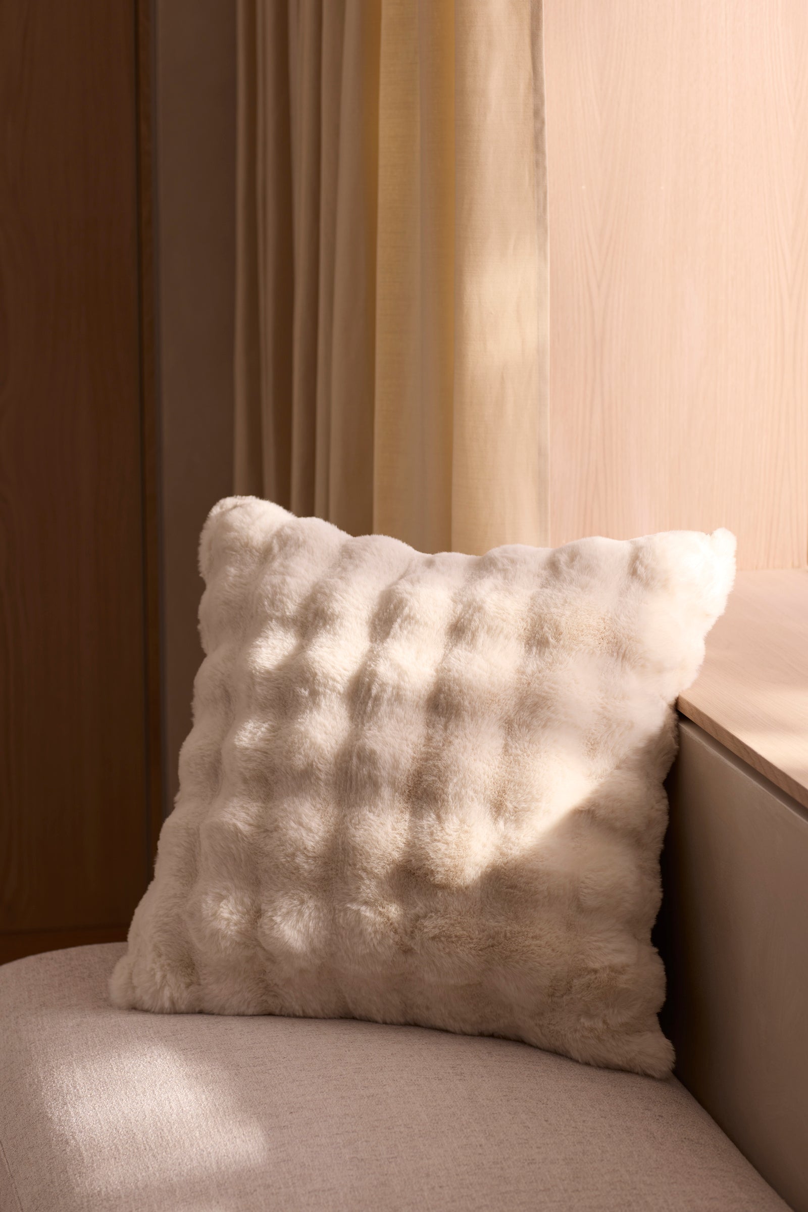 The Bubble Cuddle Pillow by Cozy Earth, soft and white with a textured design, sits on a light sofa beside a wooden wall and beige curtains, bathed in natural sunlight. 