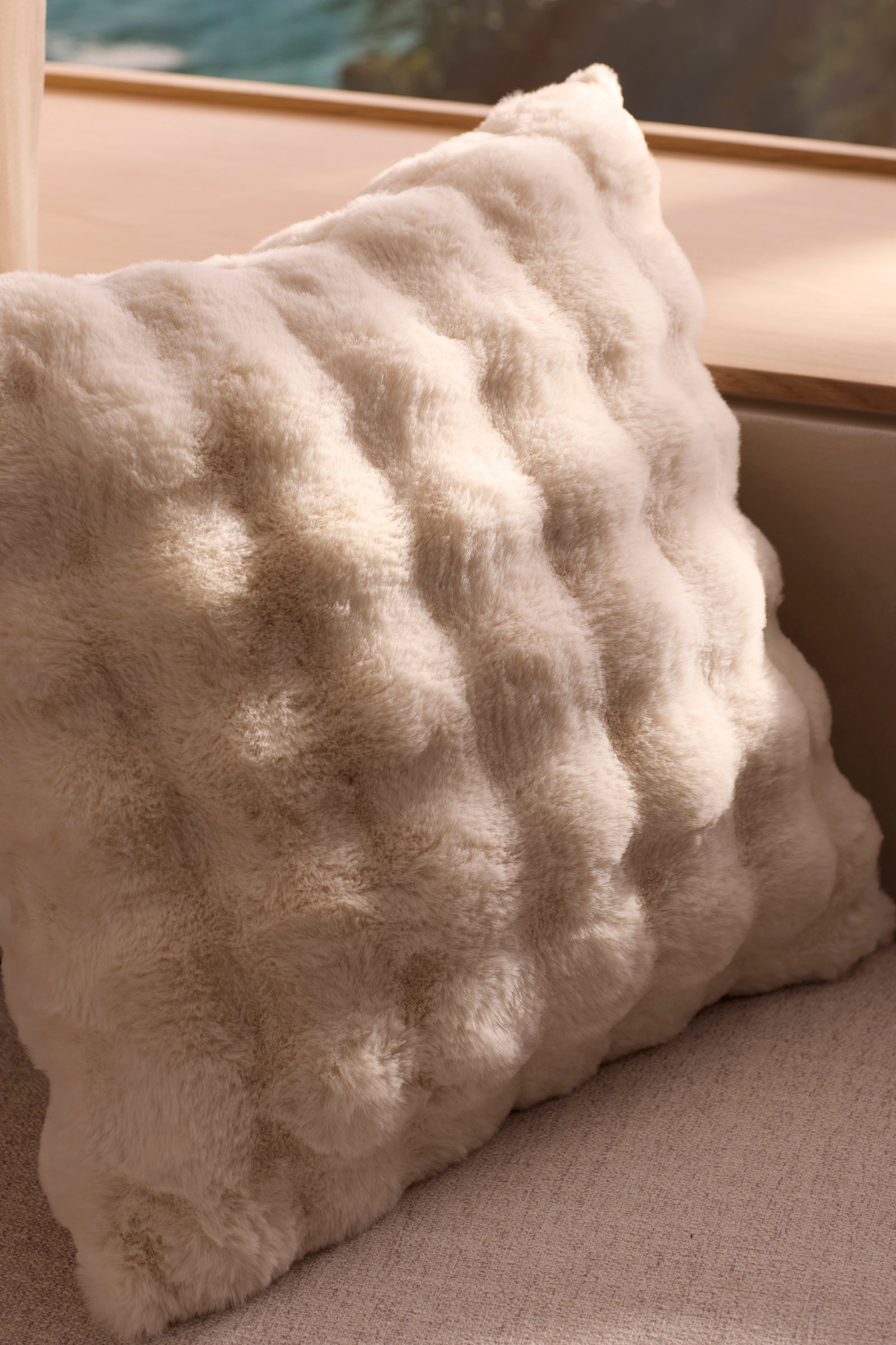 The Bubble Cuddle Pillow by Cozy Earth, featuring soft cream-colored faux fur with a wavy texture, sits on a light sofa near wood. Sunlight highlights this plush, fluffy accent.