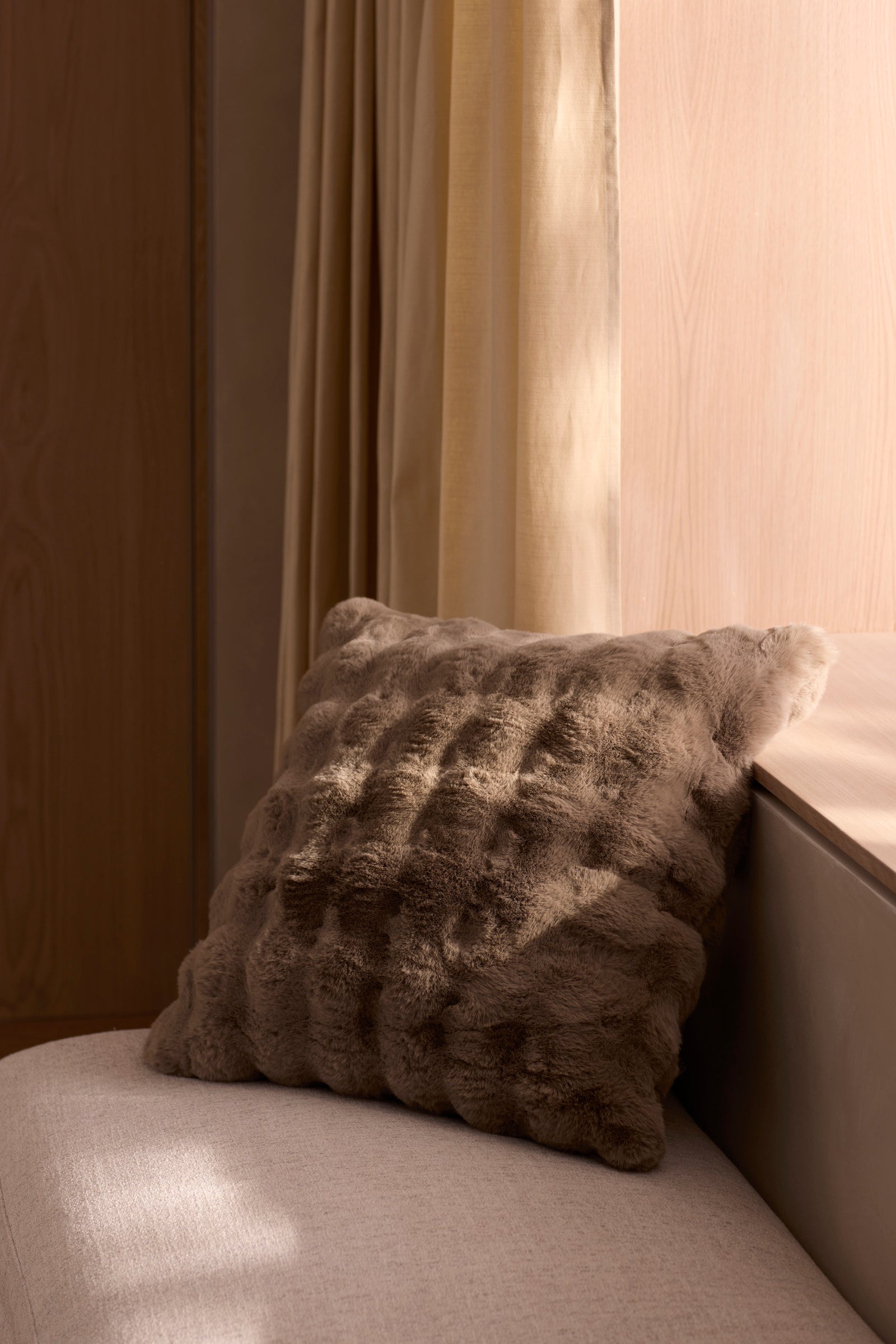 The Bubble Cuddle Pillow by Cozy Earth, featuring a soft, textured brown design, sits on a light cushioned bench beside a wooden wall and window draped in beige curtains, all bathed in natural sunlight. 