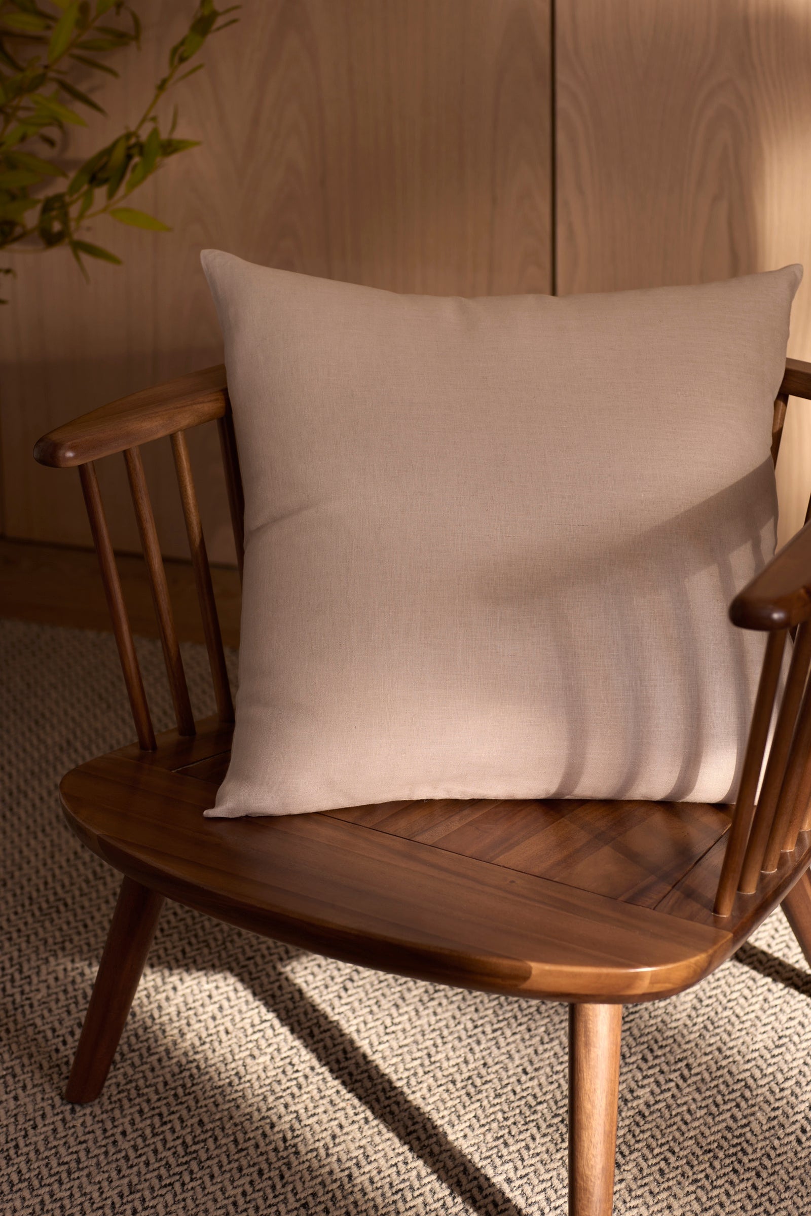 A Cozy Earth Linen Throw Pillow rests on a wooden chair with a light beige cushion atop a textured woven rug, as sunlight casts soft shadows and a green plant peeks in the background. 