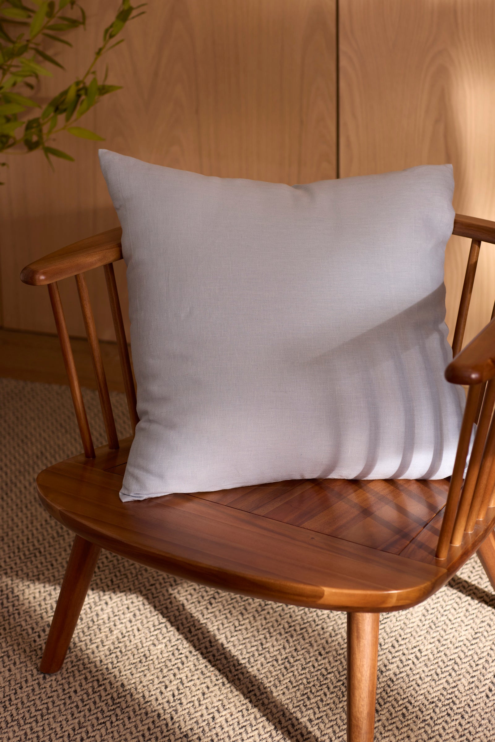 The Cozy Earth Linen Throw Pillow sits on a wooden chair with a spindle backrest, atop a patterned rug in a room featuring light wood paneling and a plant in the background.
