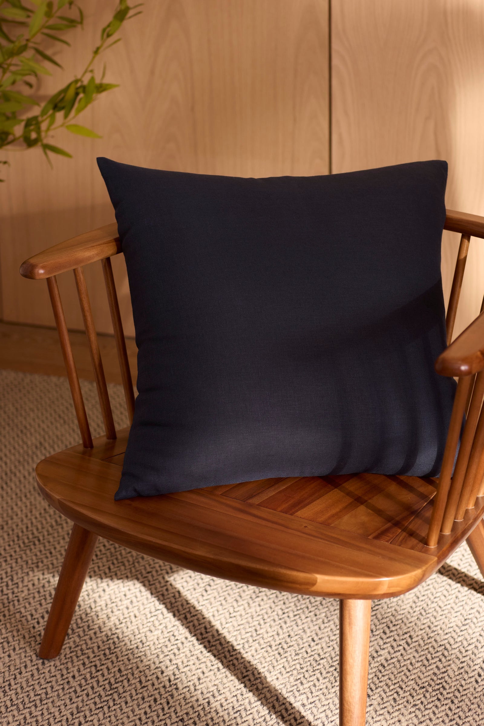 The Cozy Earth Linen Throw Pillow in navy blue sits on a wooden spindle-back chair atop a textured beige rug, with a green plant and light wood paneling in the background.