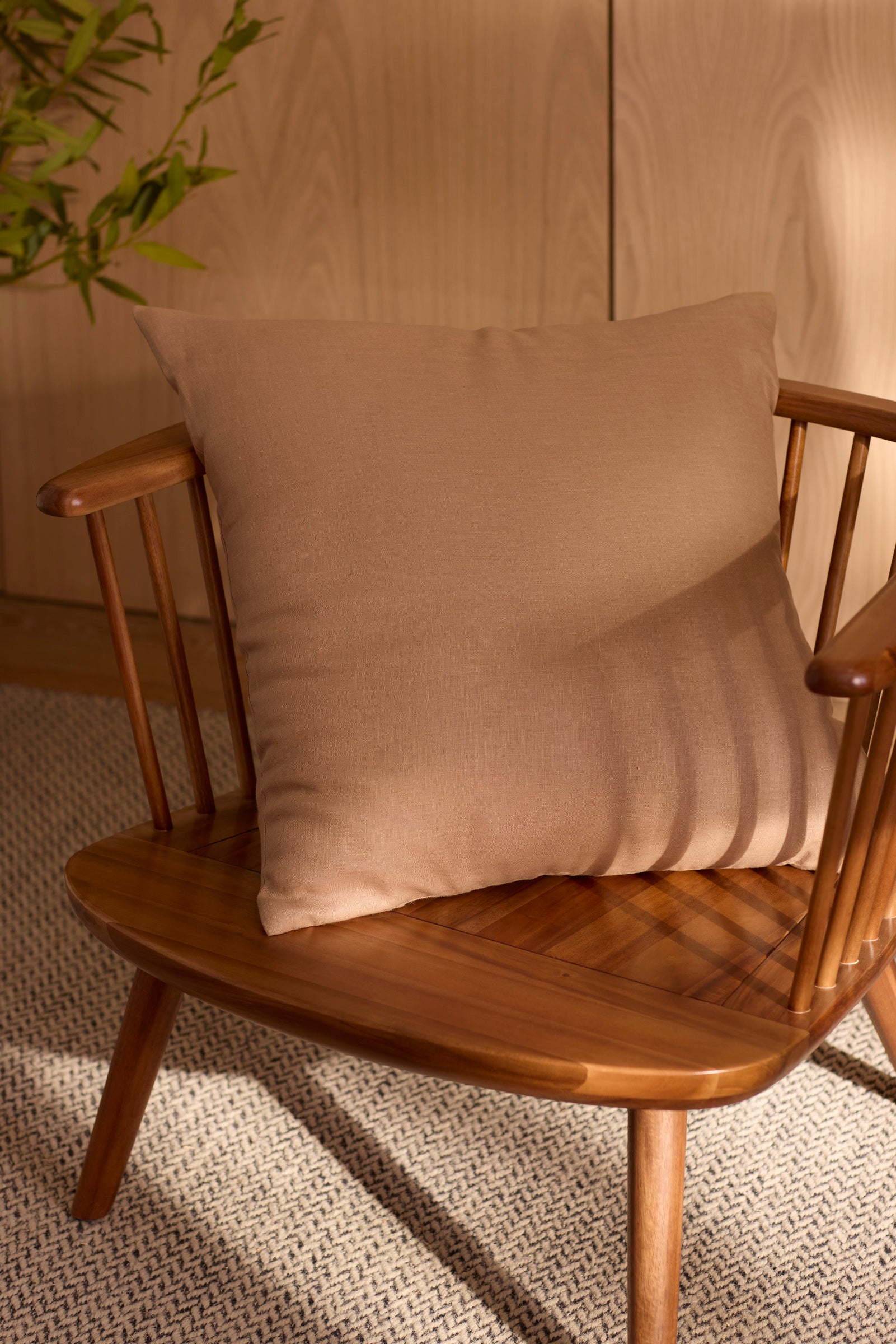 A Cozy Earth Linen Throw Pillow rests on a wooden chair with a spindle backrest, bathed in soft sunlight. A textured rug and leafy plant add coziness to the minimalist space.