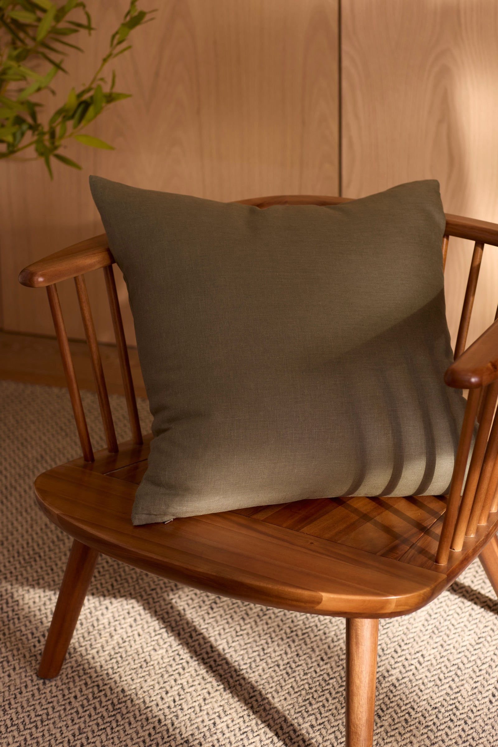 A wooden chair with vertical slats holds a Cozy Earth Linen Throw Pillow in dark green. The chair sits on textured carpet as soft sunlight casts shadows, with a green plant partially visible in the background. 