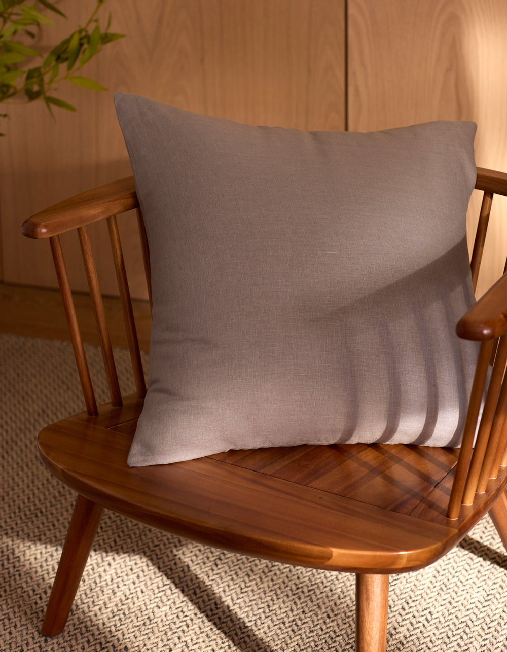 The Cozy Earth Linen Throw Pillow in light gray rests on a wooden spindle-back chair. Sunlight creates soft shadows, while a woven rug and green plant enhance the inviting background.