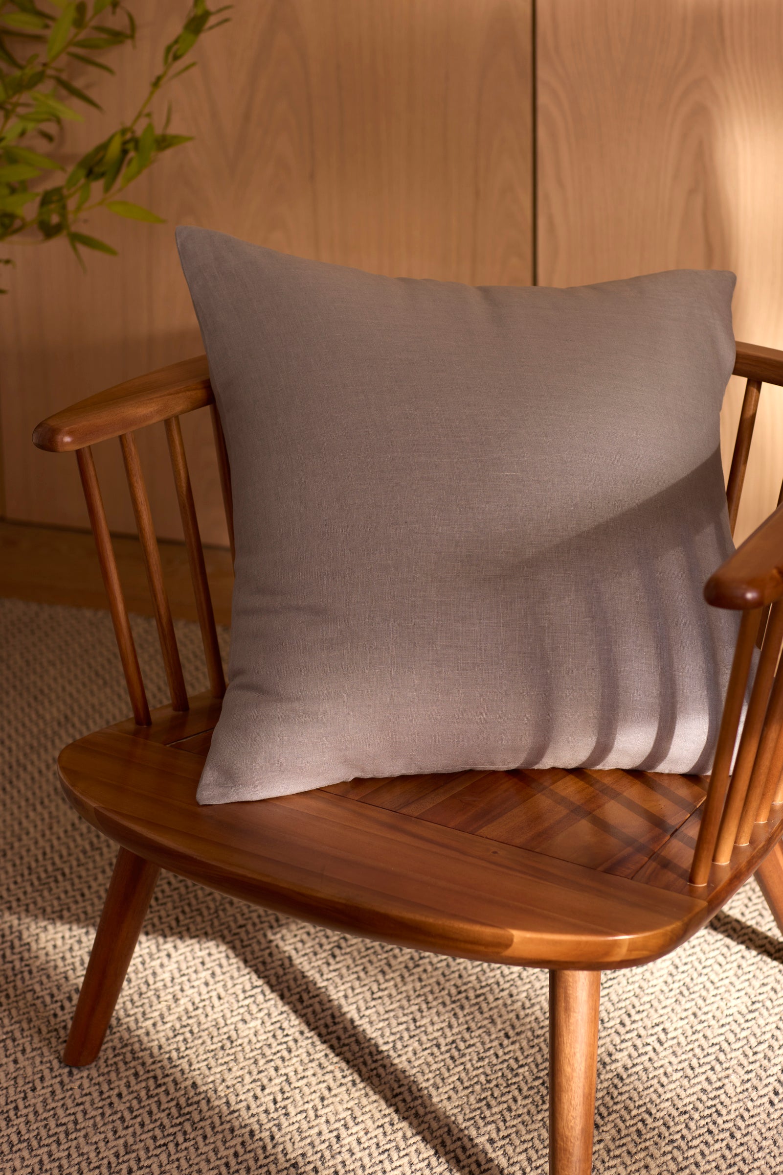 A Linen Throw Pillow by Cozy Earth rests on a wooden chair beside a woven rug, sunlight highlighting its texture. In the background, a leafy green plant and part of a wooden cabinet are visible. 