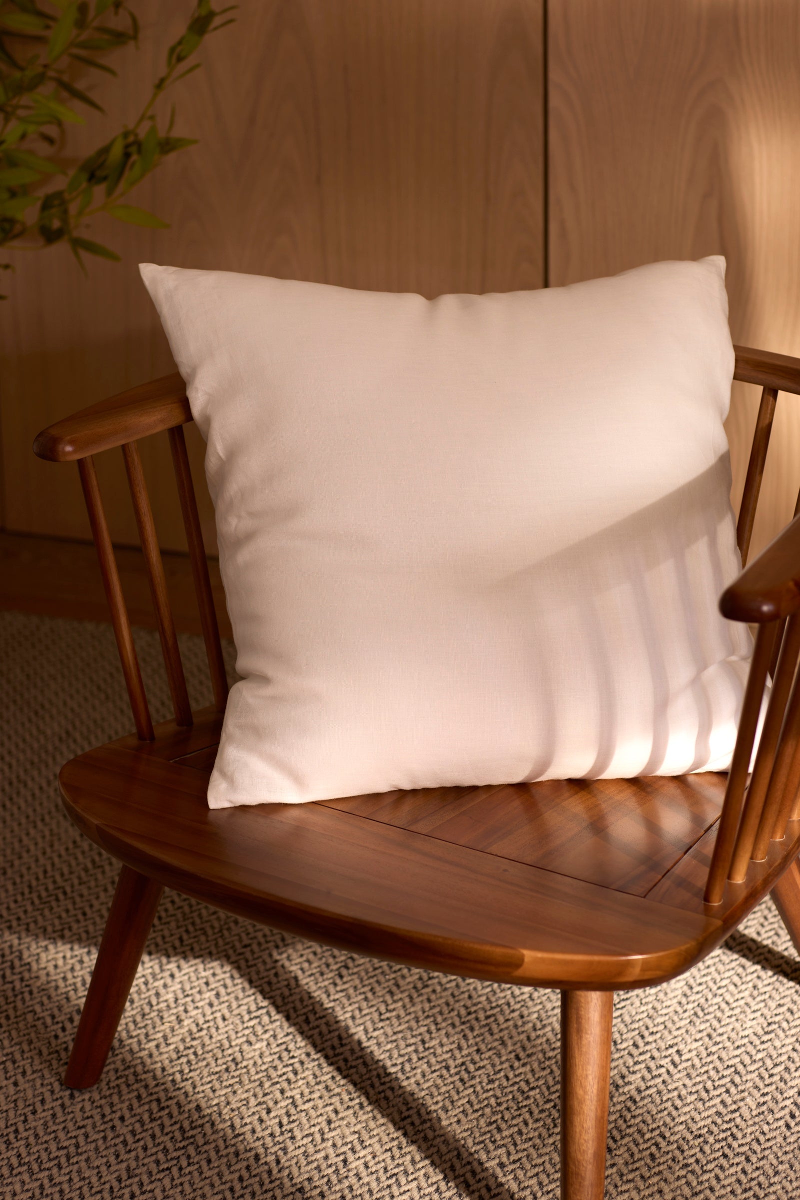 A Cozy Earth Linen Throw Pillow sits on a wooden chair with vertical slats, placed on a textured rug. Sunlight highlights the chair, with a wooden cabinet and green plant nearby. 