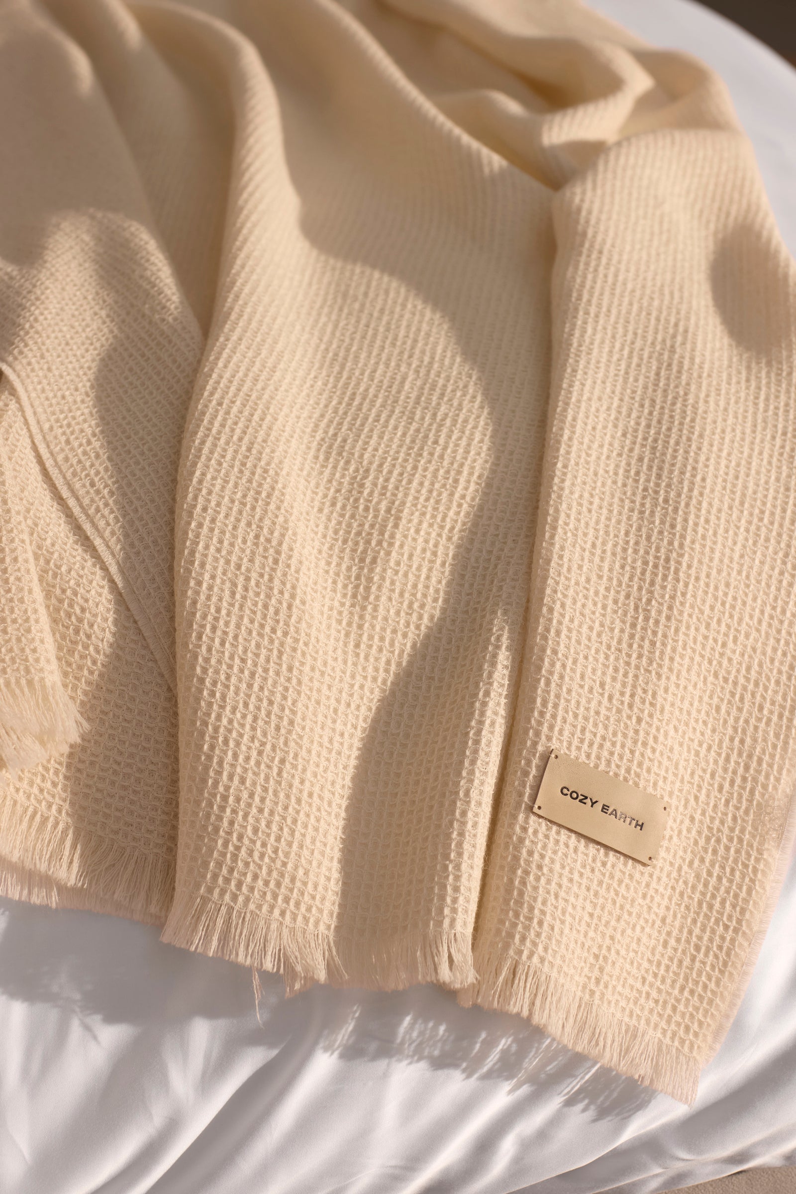 The Cozy Earth Alpaca Waffle Throw features a beige, textured waffle weave and fringed edges. A small "Cozy Earth" tag is attached, and the soft throw is displayed on a white surface. 