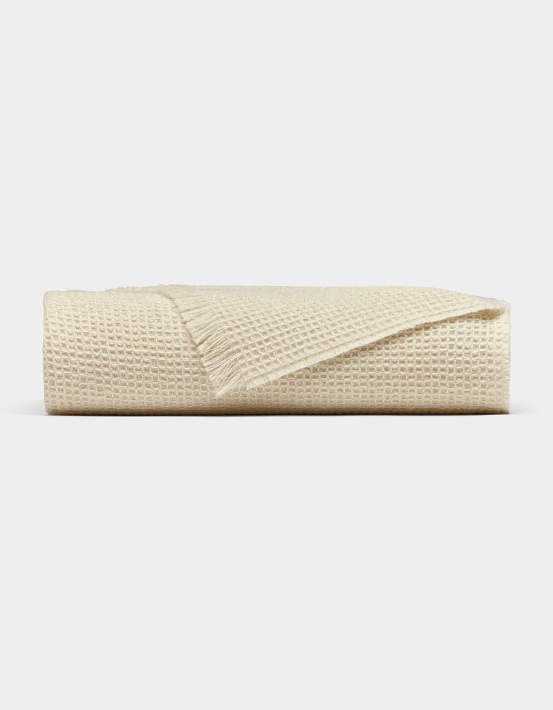 The Cozy Earth Alpaca Waffle Throw, in beige with a fringed edge, is neatly folded and displayed against a plain light gray background.