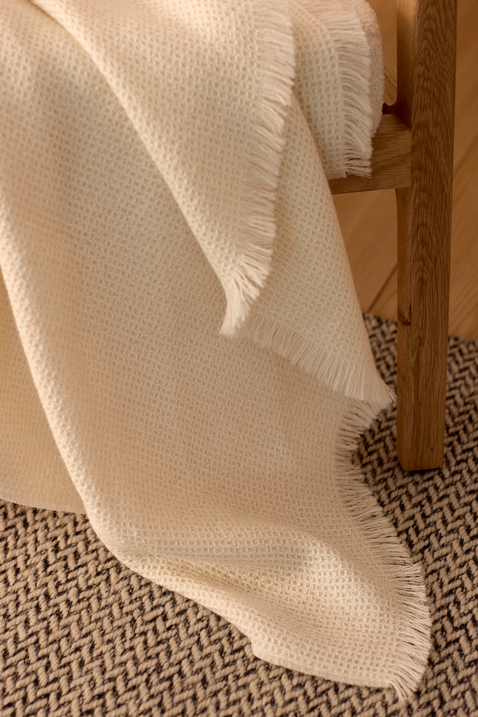 The Cozy Earth Alpaca Waffle Throw, in a cream hue with textured weave and fringed edges, is draped over a wooden chair and cascades onto a black-and-white patterned rug on a wooden floor. 