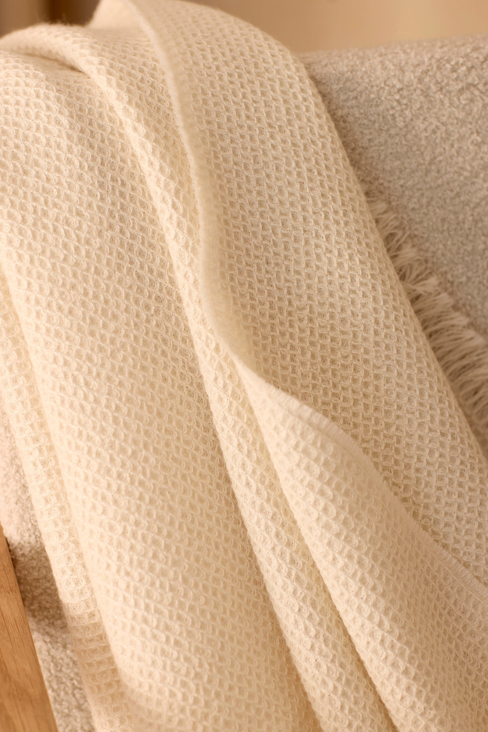 A close-up of the Alpaca Waffle Throw by Cozy Earth, featuring an off-white textured waffle-weave, draped casually over a beige soft fabric surface. 