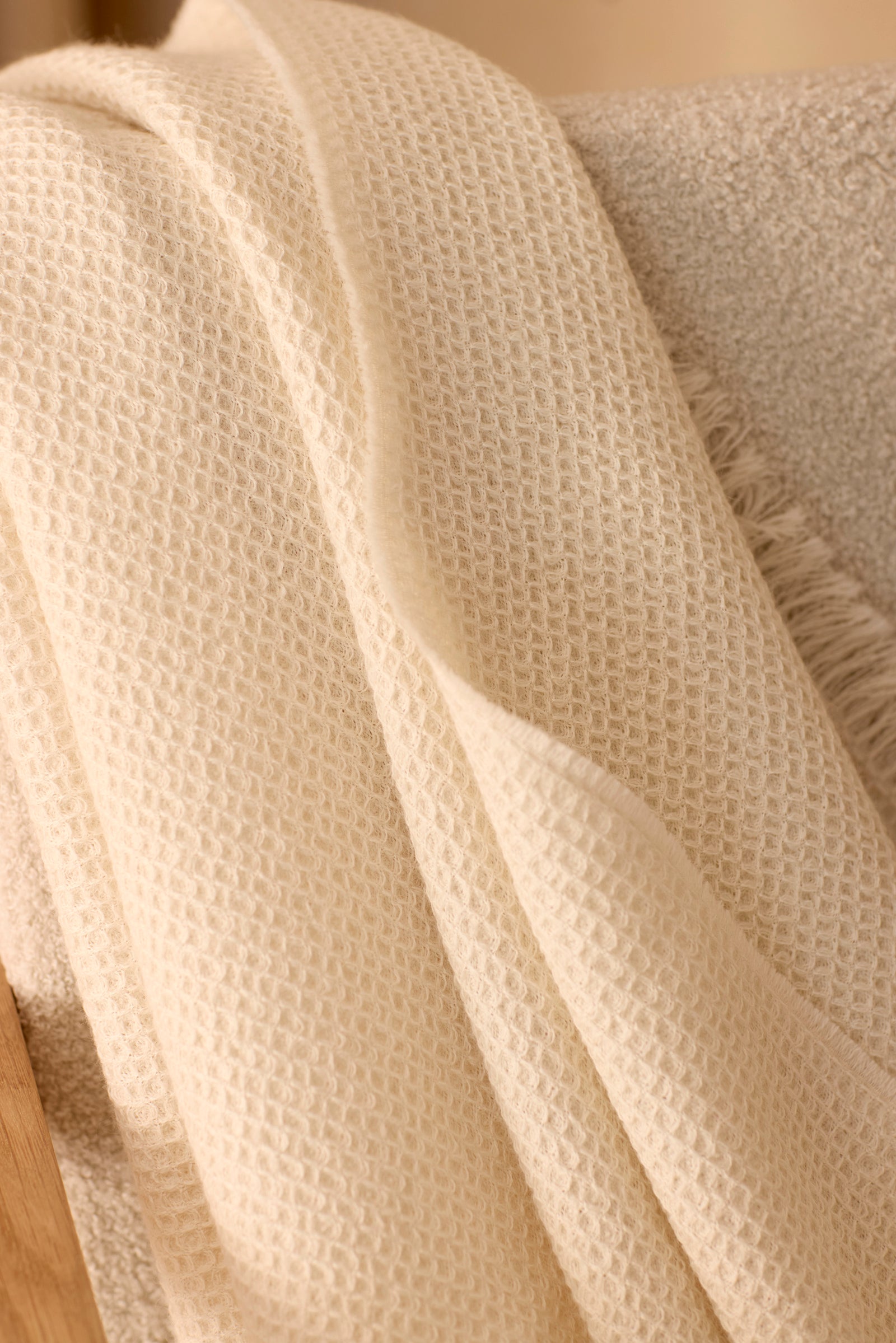 A close-up of the Alpaca Waffle Throw by Cozy Earth, featuring an off-white textured waffle-weave, draped casually over a beige soft fabric surface. 