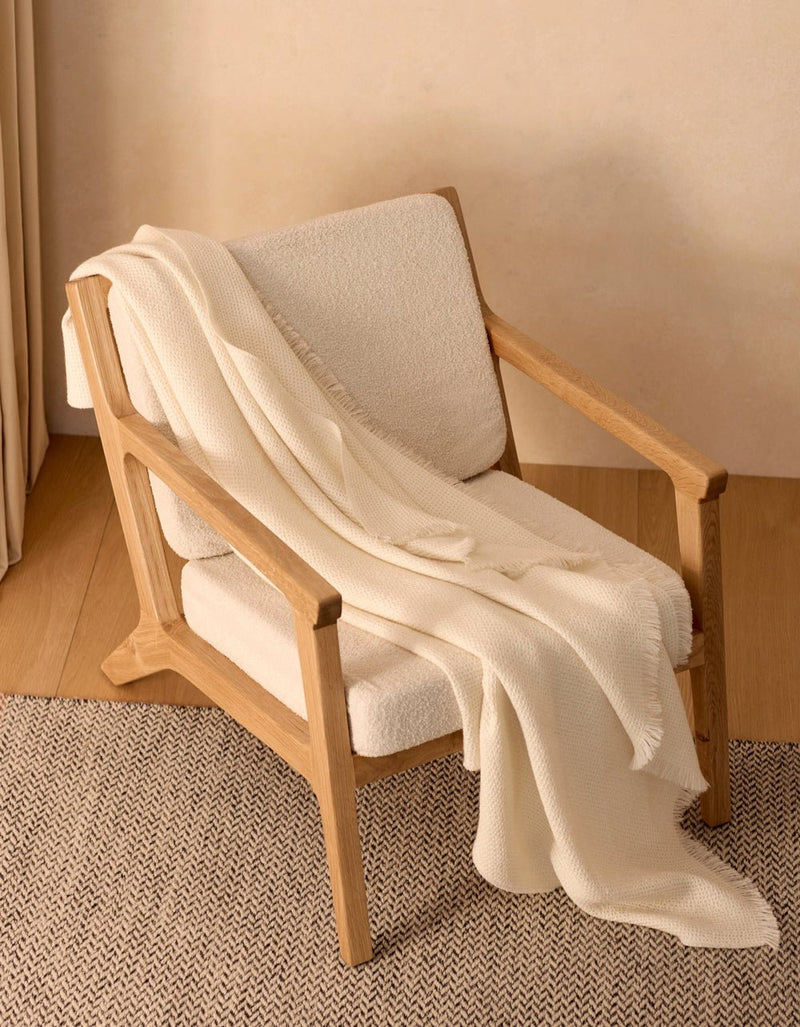 A modern wooden armchair with light beige upholstery sits on a herringbone rug, draped with the Cozy Earth Alpaca Waffle Throw. The soft blanket and minimal setting create a warm, inviting atmosphere.
