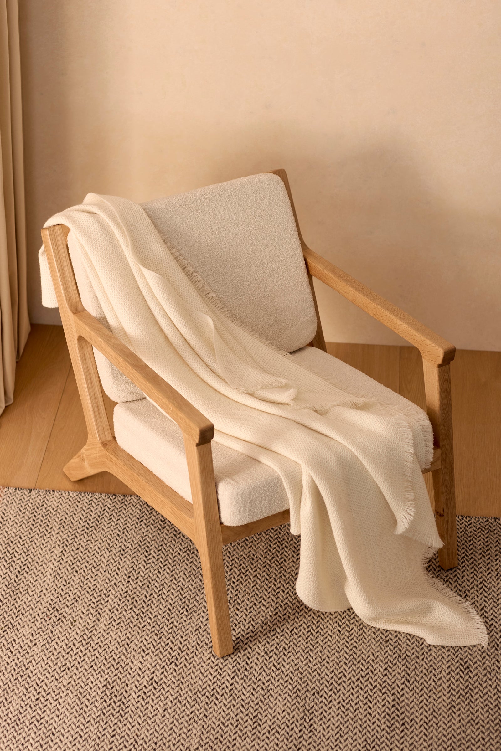 A light wooden armchair with cream-colored cushions sits on a beige carpet, draped with the Cozy Earth Alpaca Waffle Throw. The scene is minimal and cozy, featuring soft neutrals. 