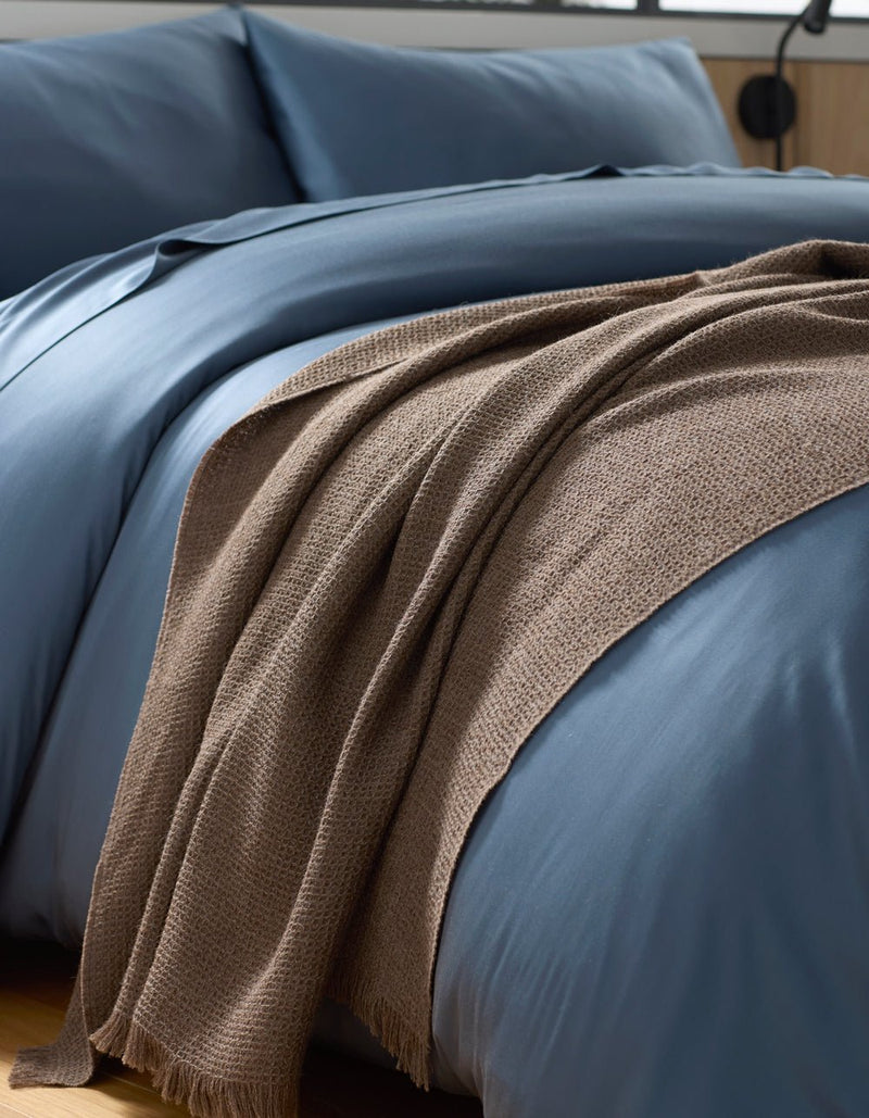 A close-up of a neatly made bed with smooth blue bedding and a Cozy Earth Alpaca Waffle Throw in brown draped casually over the edge.