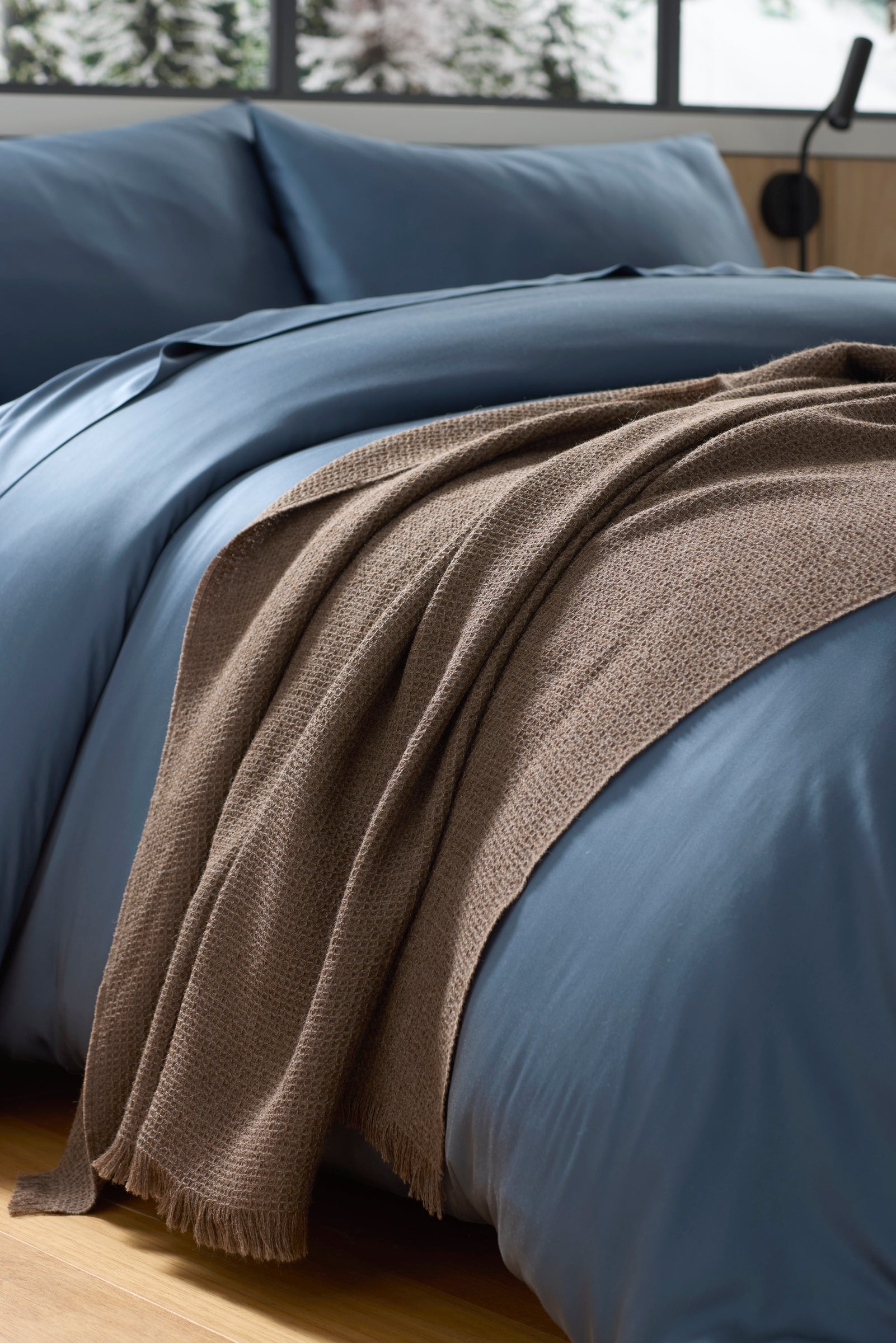 A close-up of a neatly made bed with smooth blue bedding, accented by the Cozy Earth Alpaca Waffle Throw in brown, draped across it. A portion of a window reveals a snowy outdoor scene in the background. |Color:Ebony
