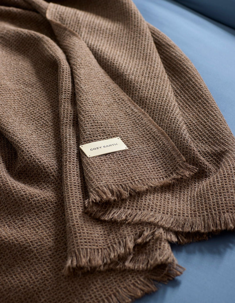 A brown Alpaca Waffle Throw by Cozy Earth, featuring a textured weave and fringed edges, is draped over a blue surface. A small white label with the brand name "Cozy Earth" is visible on the blanket.