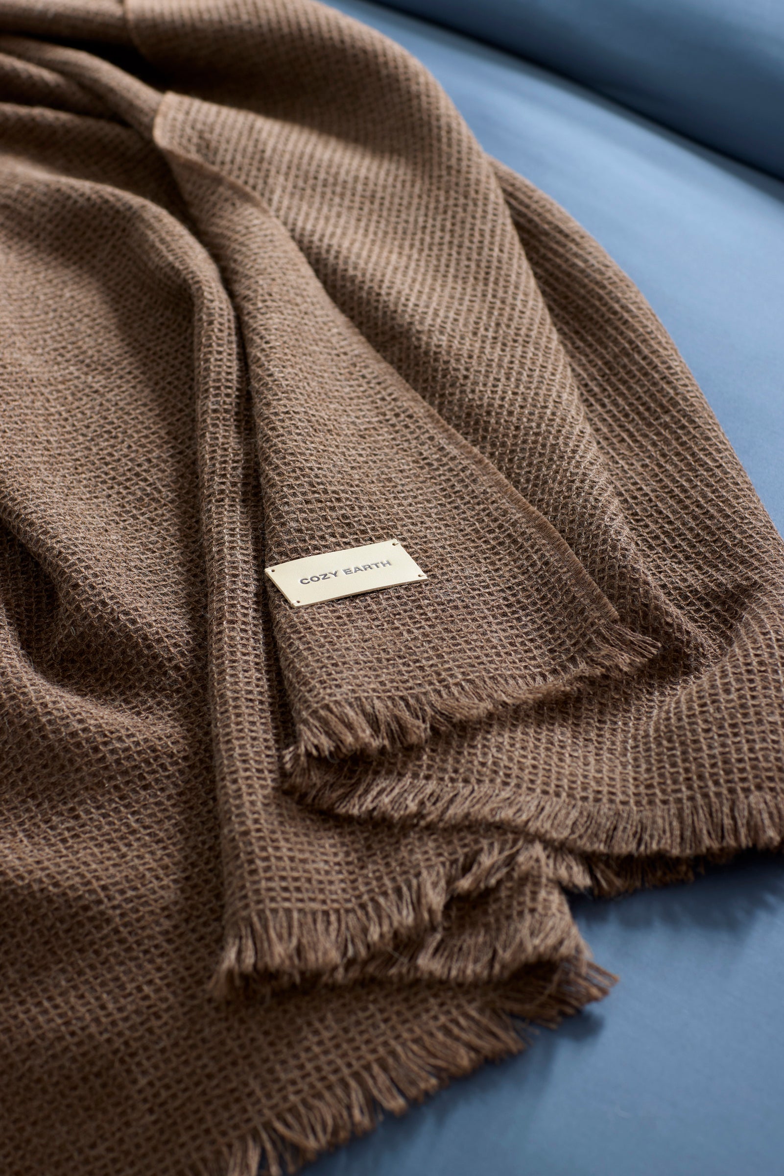 The Alpaca Waffle Throw by Cozy Earth, a textured brown blanket with fringed edges and a sewn "COZY EARTH" label, is draped over a blue surface. |Color:Ebony