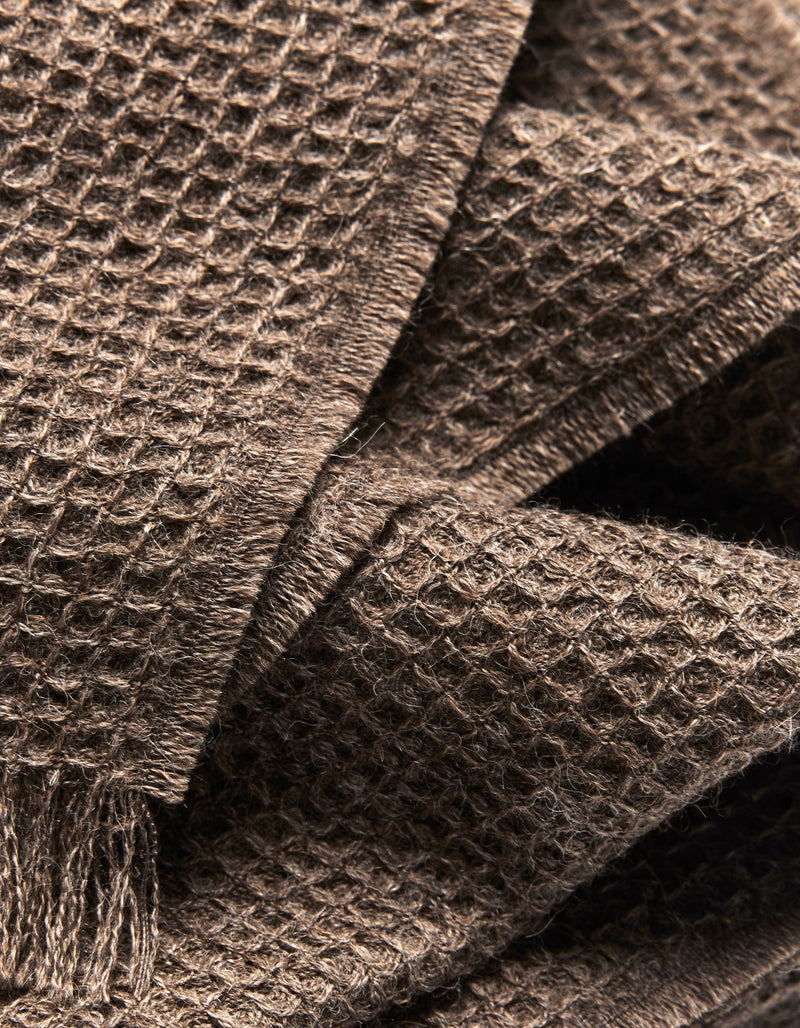 Close-up of the Alpaca Waffle Throw by Cozy Earth, highlighting its brown waffle weave texture, soft fibers, and layered folds. Fringed edges are visible in the lower left corner, emphasizing its cozy appeal.