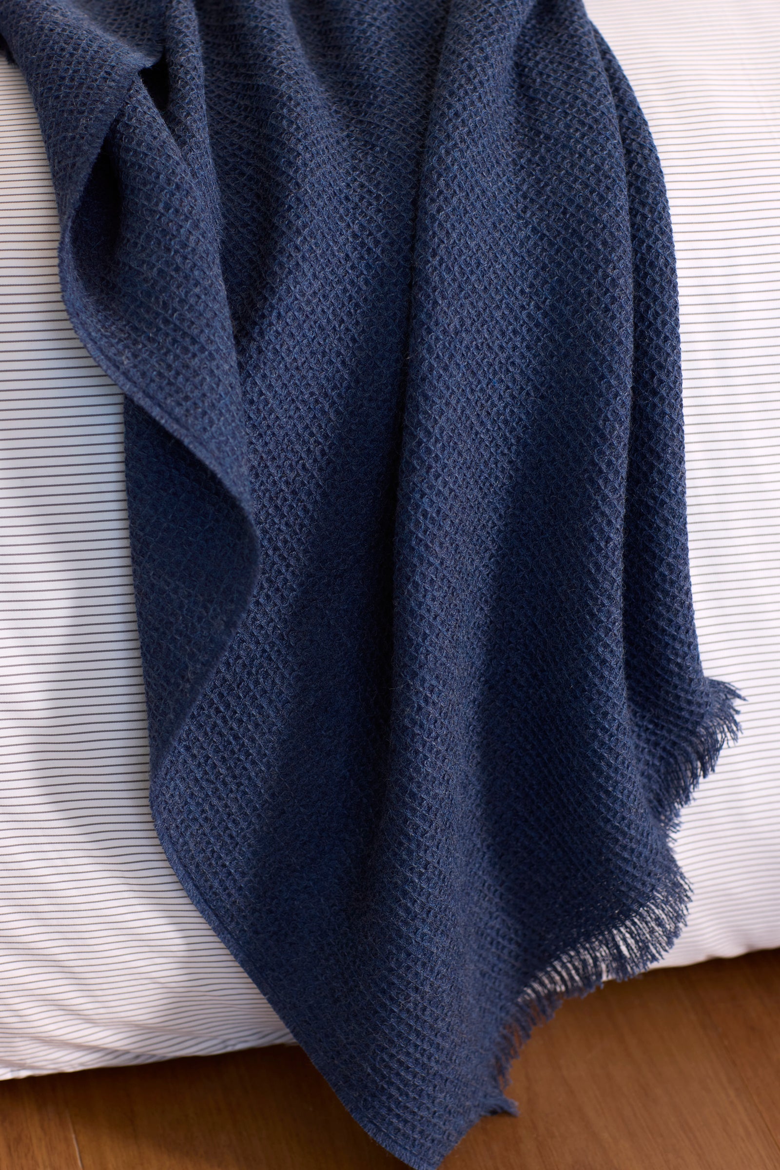 The Cozy Earth Alpaca Waffle Throw, a textured navy blue blanket with fringed edges, is draped over a white striped cushion or mattress on a wooden floor. 