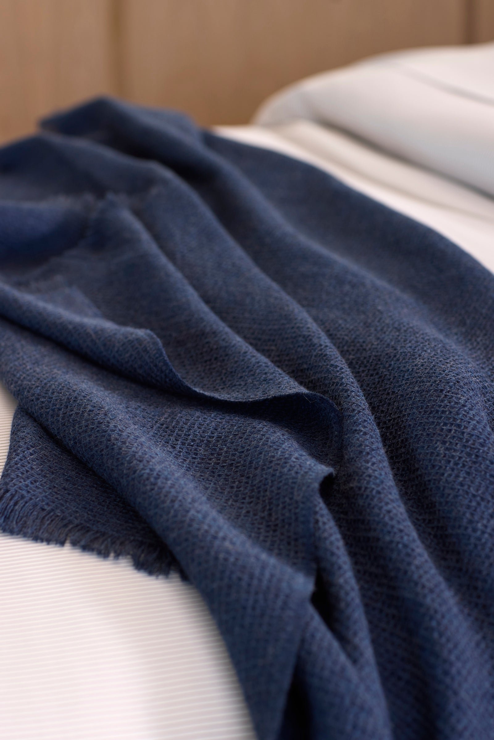 The Cozy Earth Alpaca Waffle Throw, a dark blue textured blanket with fringed edges, is draped casually over a neatly made bed with white sheets.
